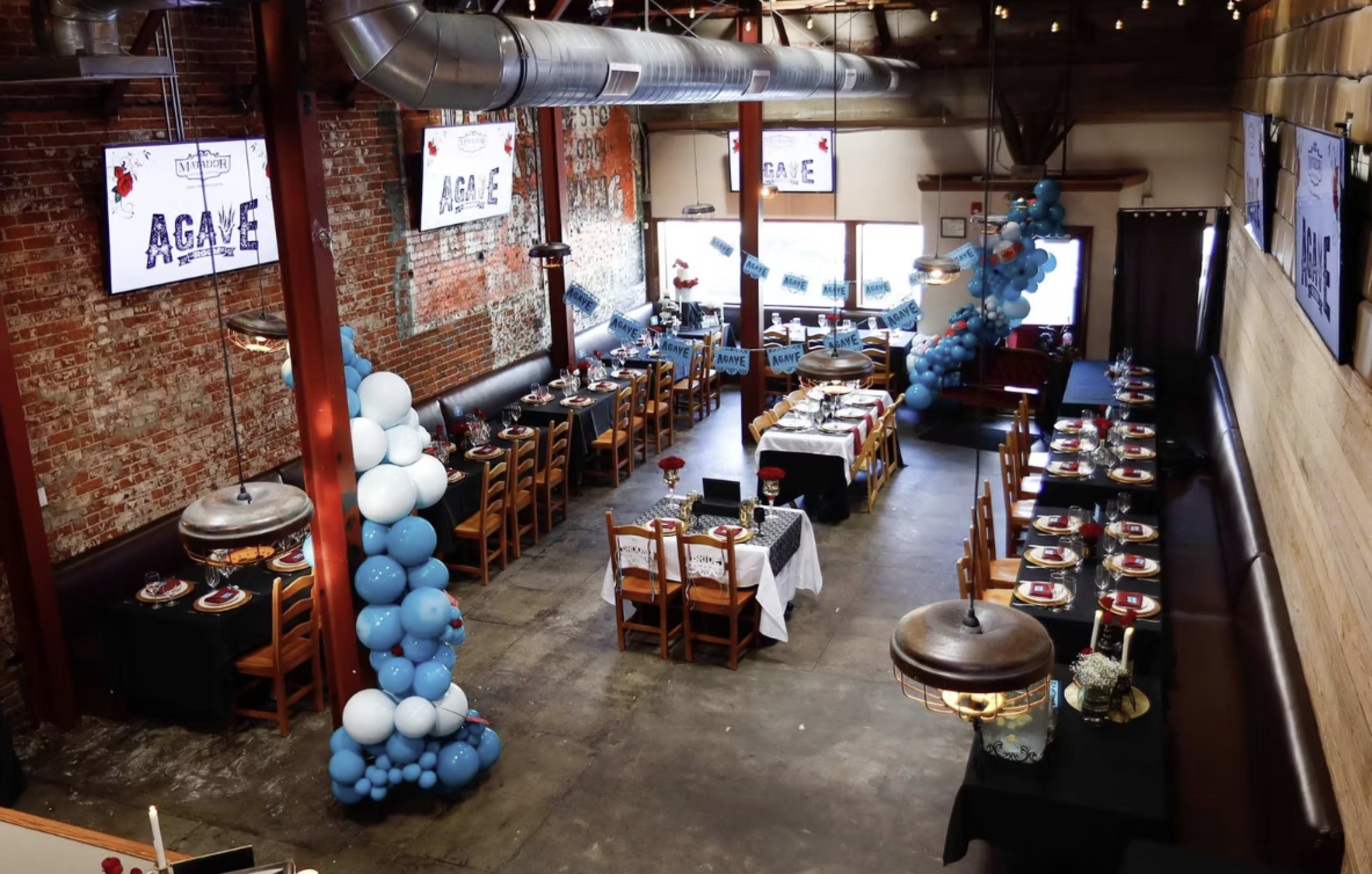 The image shows an indoor restaurant venue with long tables set for a celebration, decorated with blue balloons and large screens displaying the word "AGAVE" on the walls.