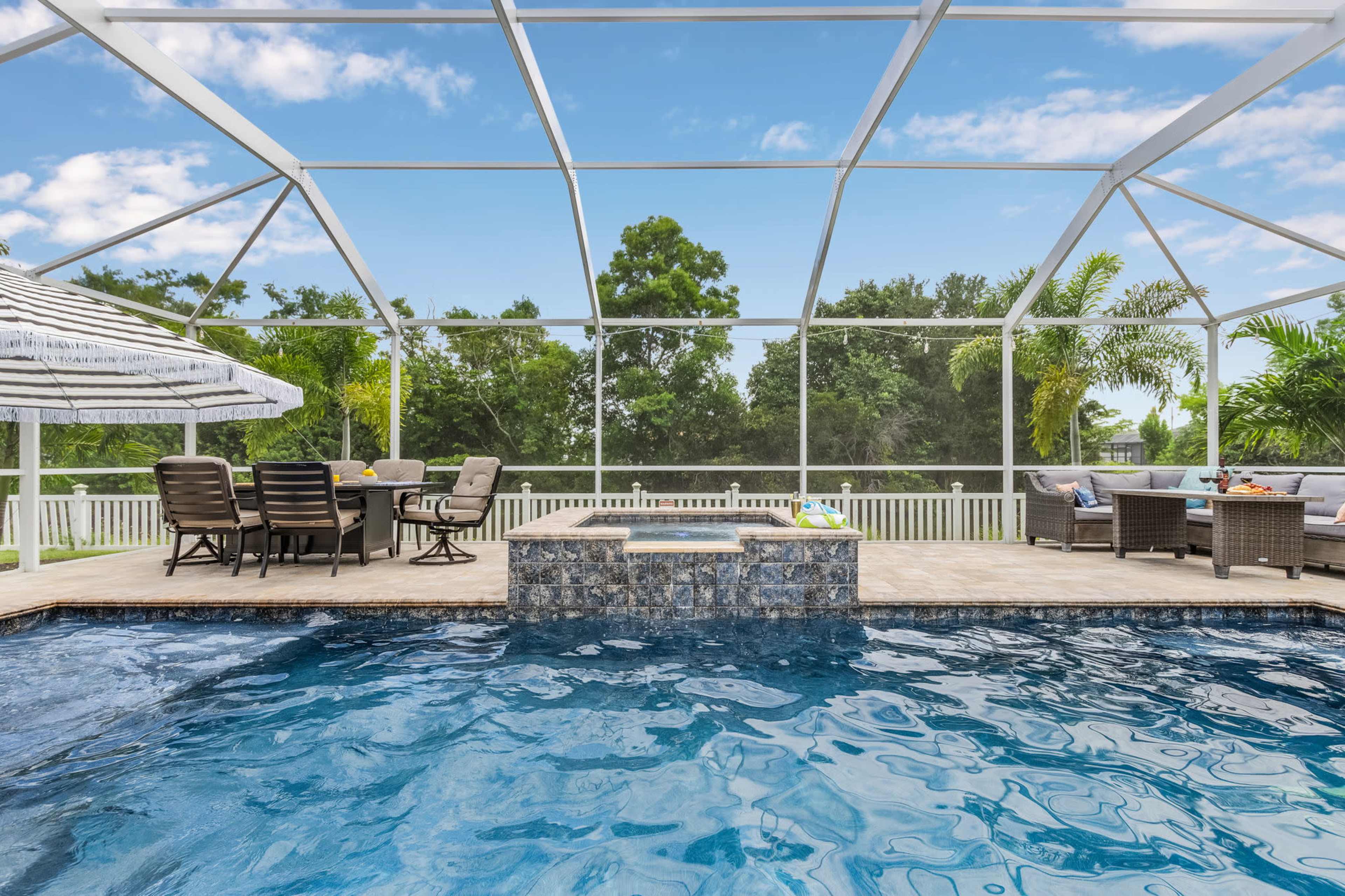 A clear pool sits within a screened patio, surrounded by lounge furniture and tropical plants against a backdrop of trees and blue skies.