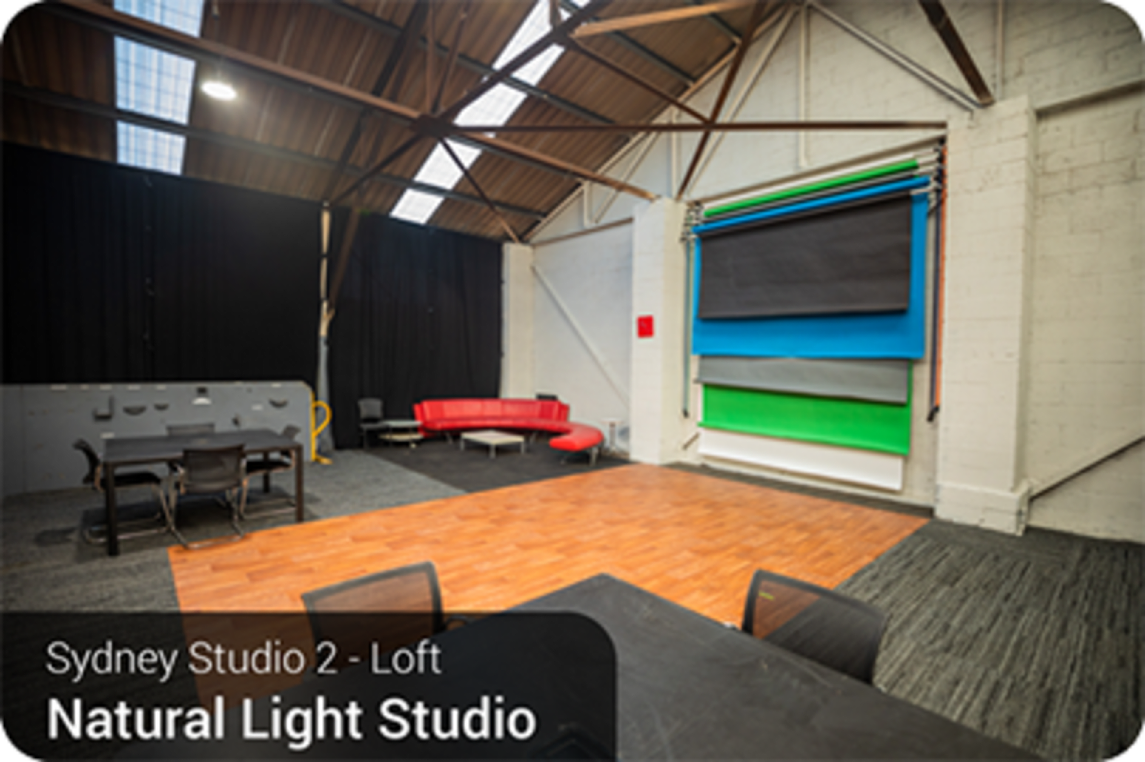 A spacious studio with a wooden floor, featuring a red seating area, several colored backdrops, and a large open ceiling with natural light.