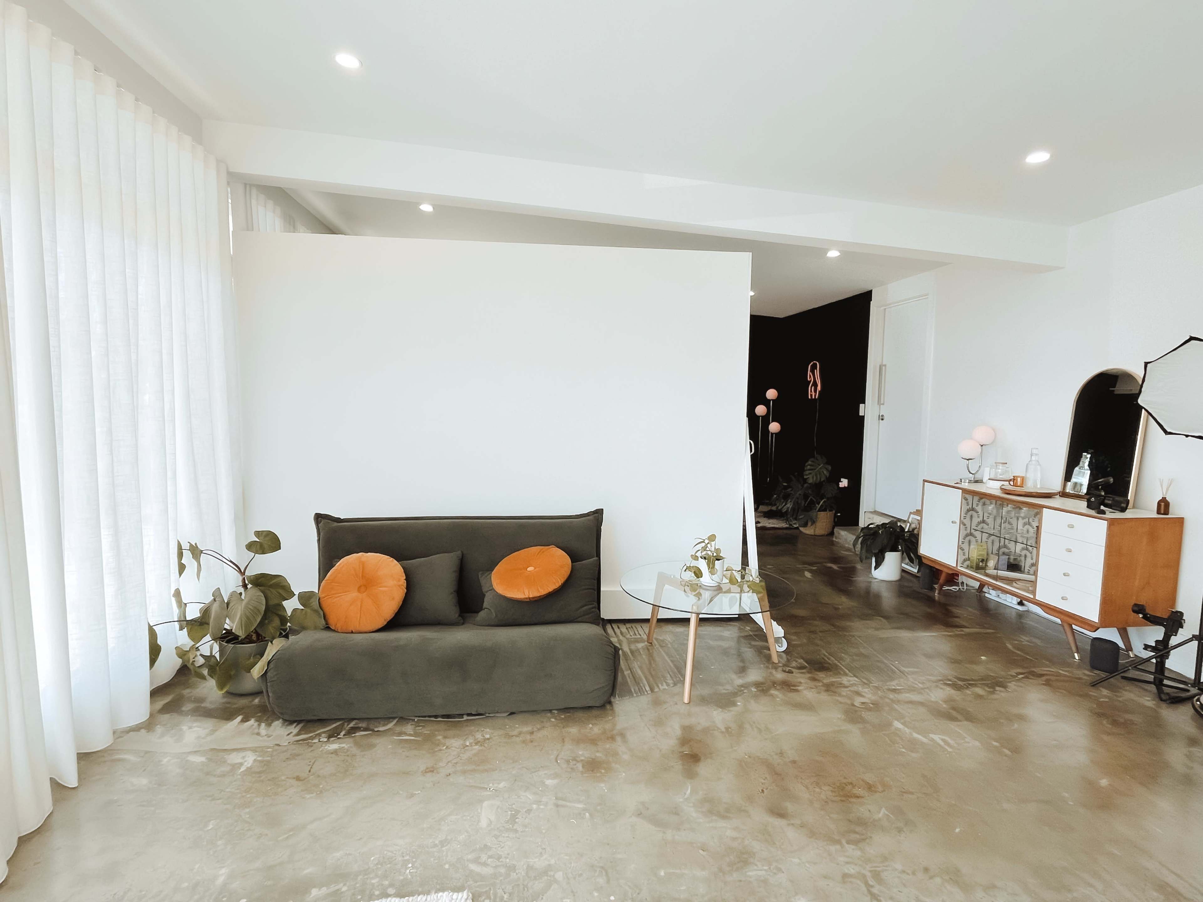 A modern living room features a dark green sofa with orange cushions, a glass coffee table, and natural light filtering through sheer curtains.