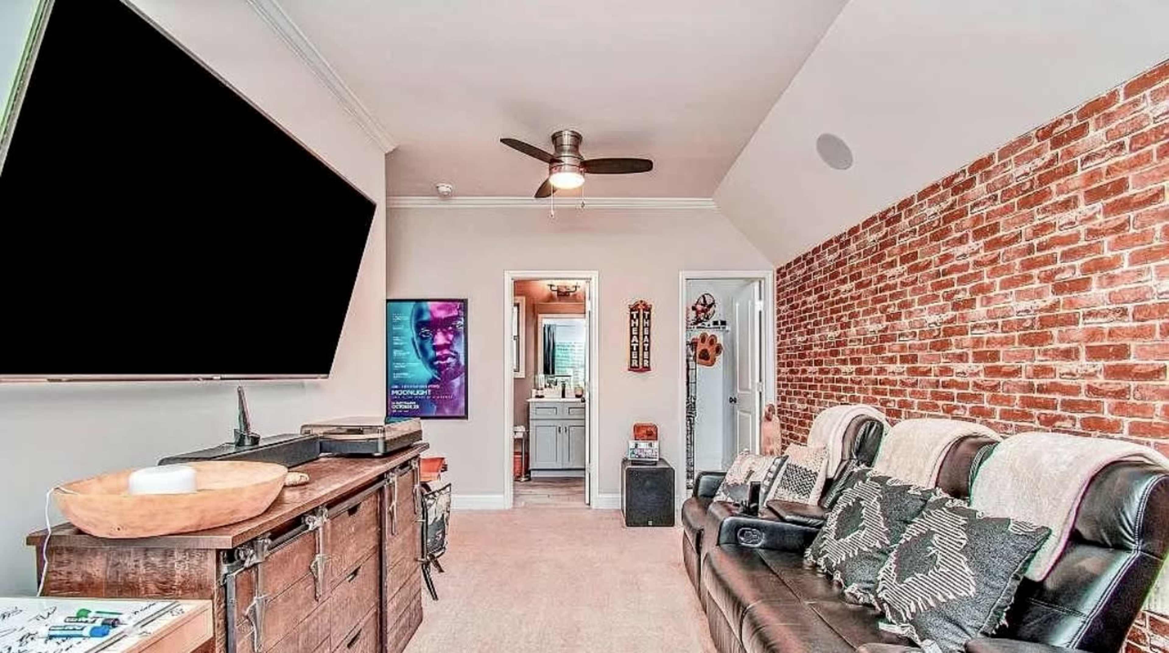 The room features a seating area with leather recliners, a brick accent wall, and an open doorway leading to a bathroom.
