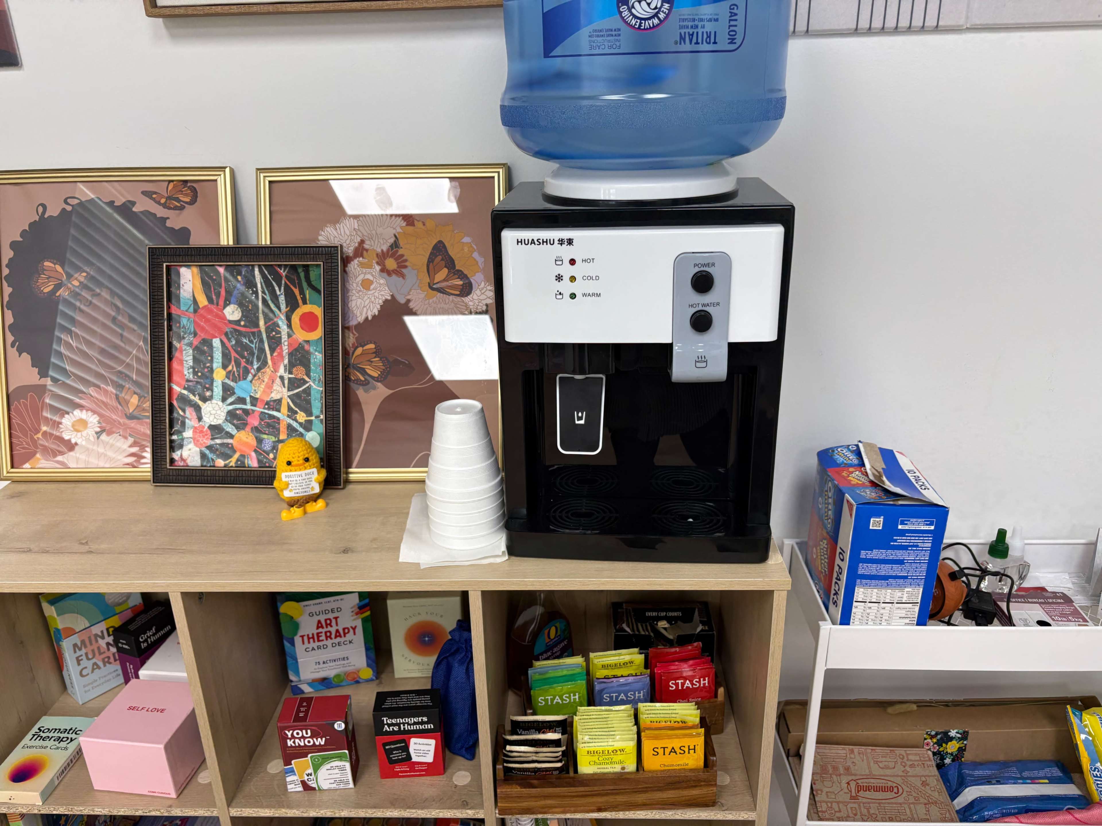 The image shows a water cooler on a wooden shelf next to framed artwork and a variety of snacks and tea bags in colorful packaging.