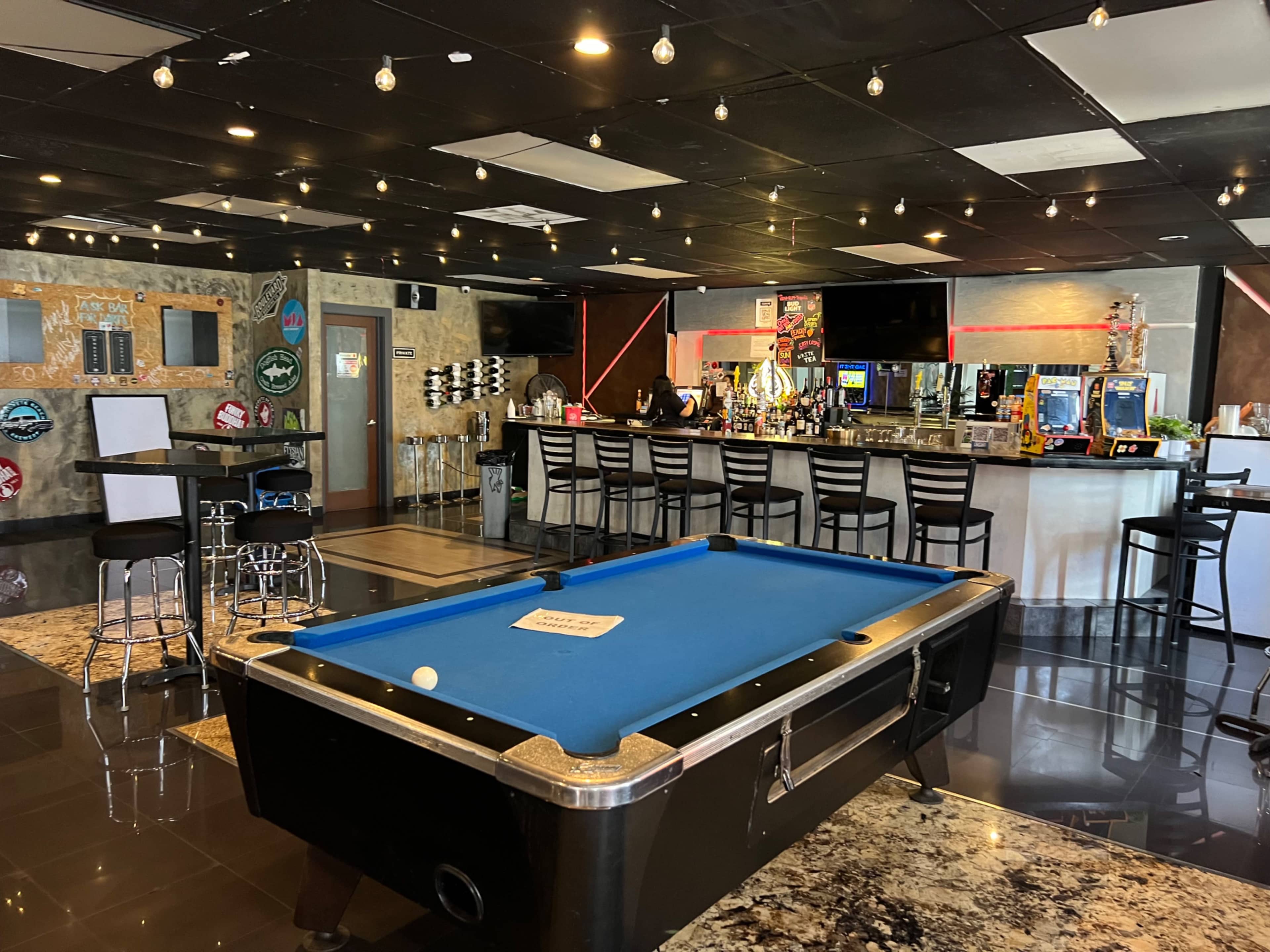 The image shows a modern bar and game room featuring a blue pool table at the center, surrounded by seating and gaming machines.