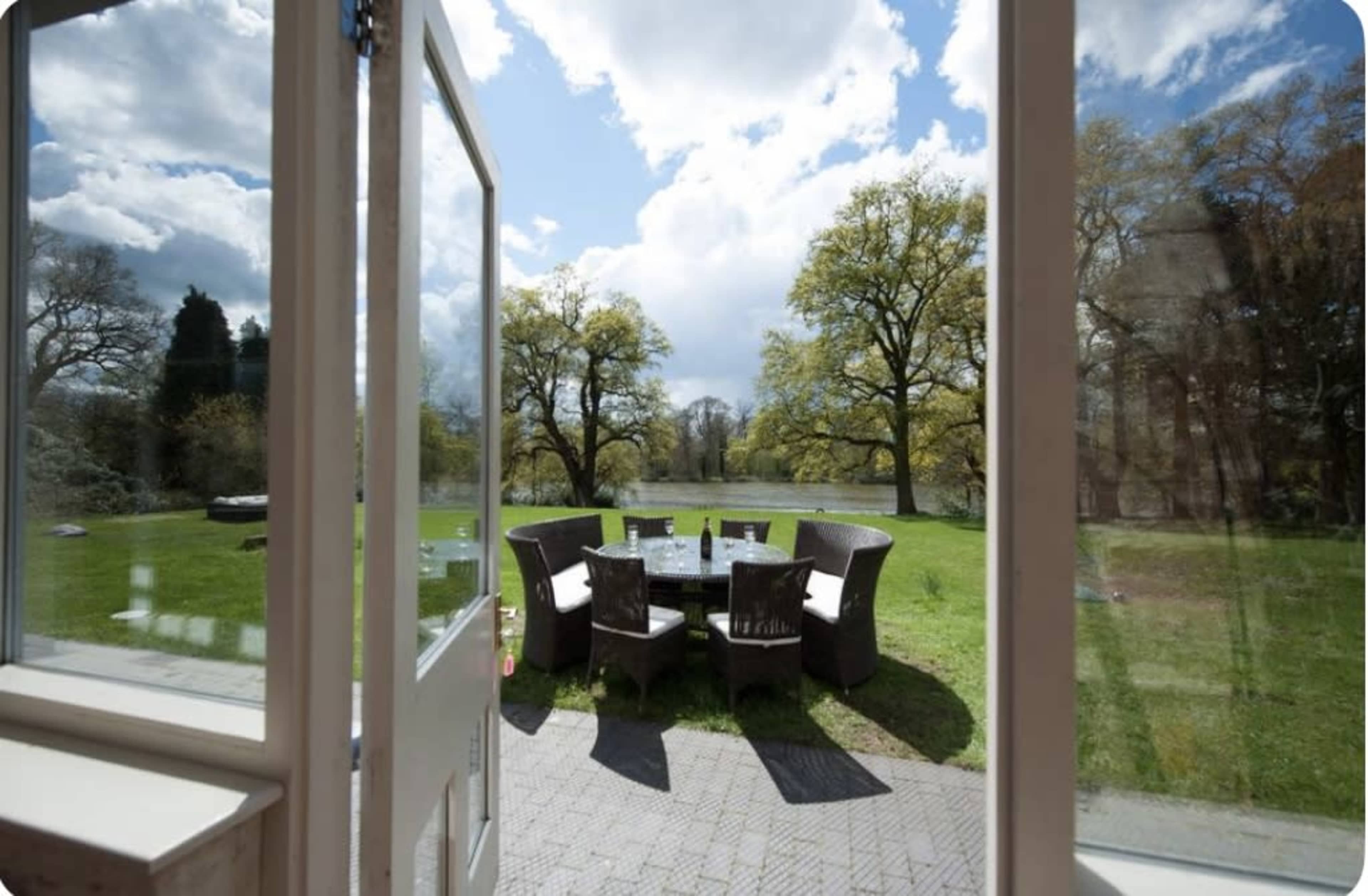 A view from an open door shows a patio with circular seating overlooking a green lawn and trees under a cloudy sky.