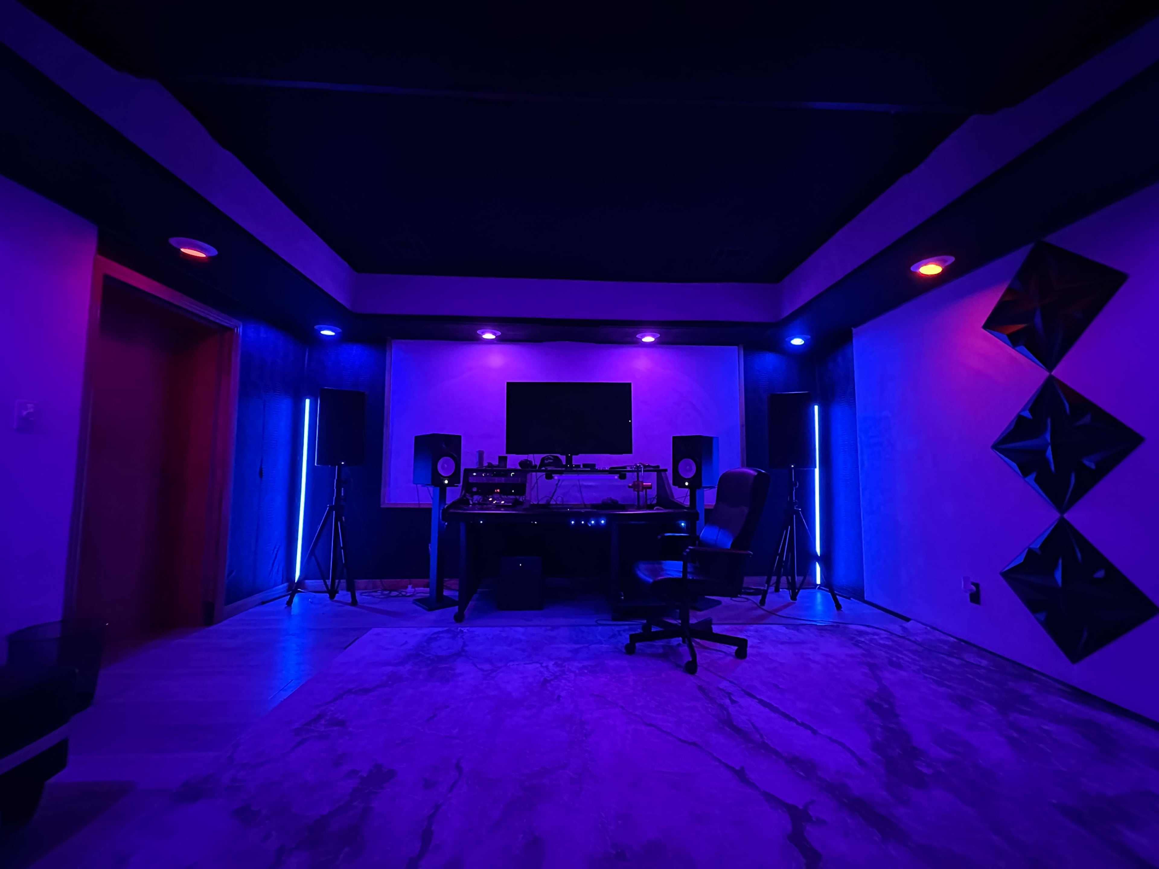 A music studio features a central mixing desk surrounded by speakers, illuminated by purple and blue lighting.