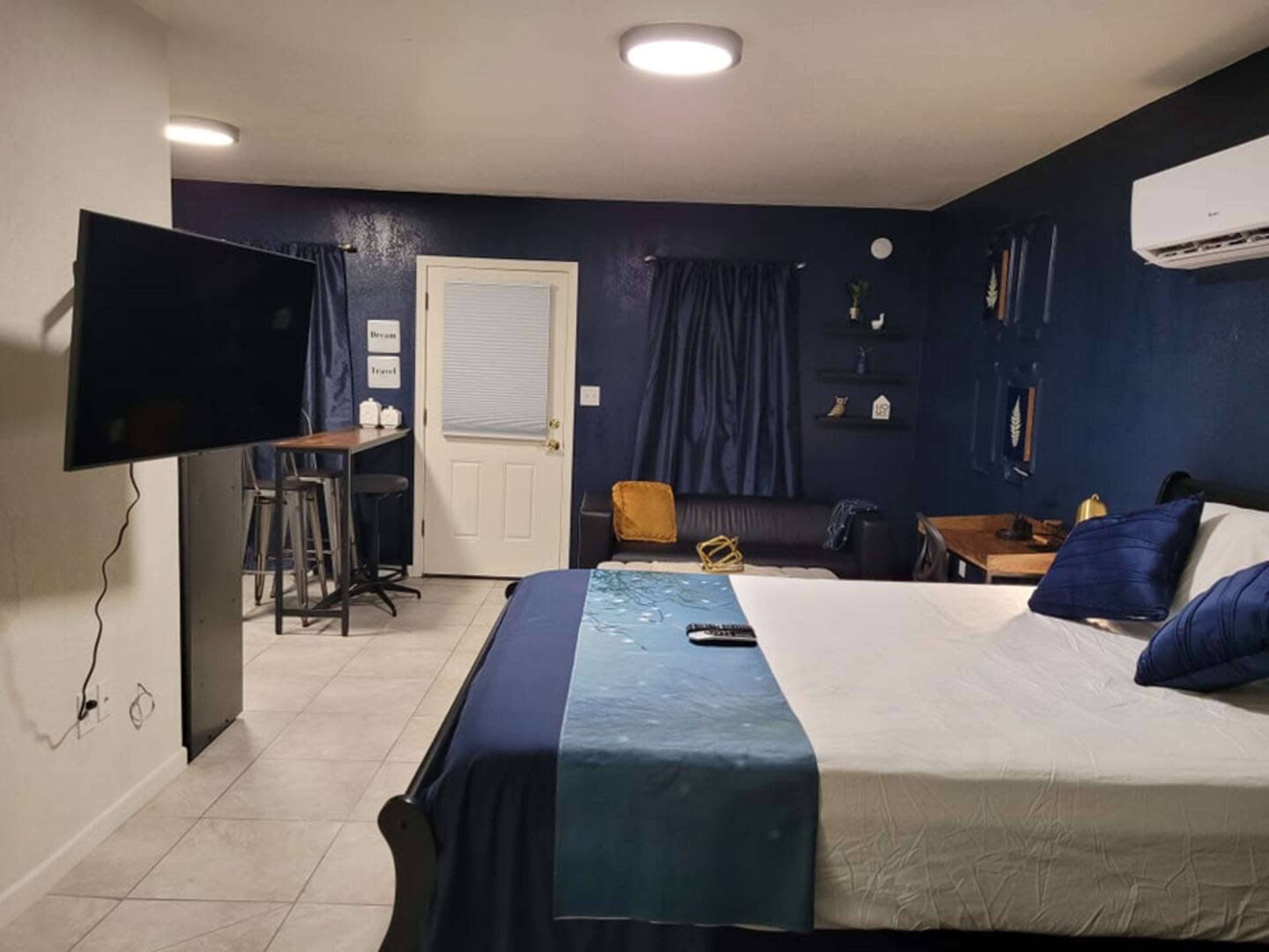 The image shows a small, modern studio apartment featuring a bed, a TV mounted on the wall, a sitting area with a couch, and a dining space with bar stools.