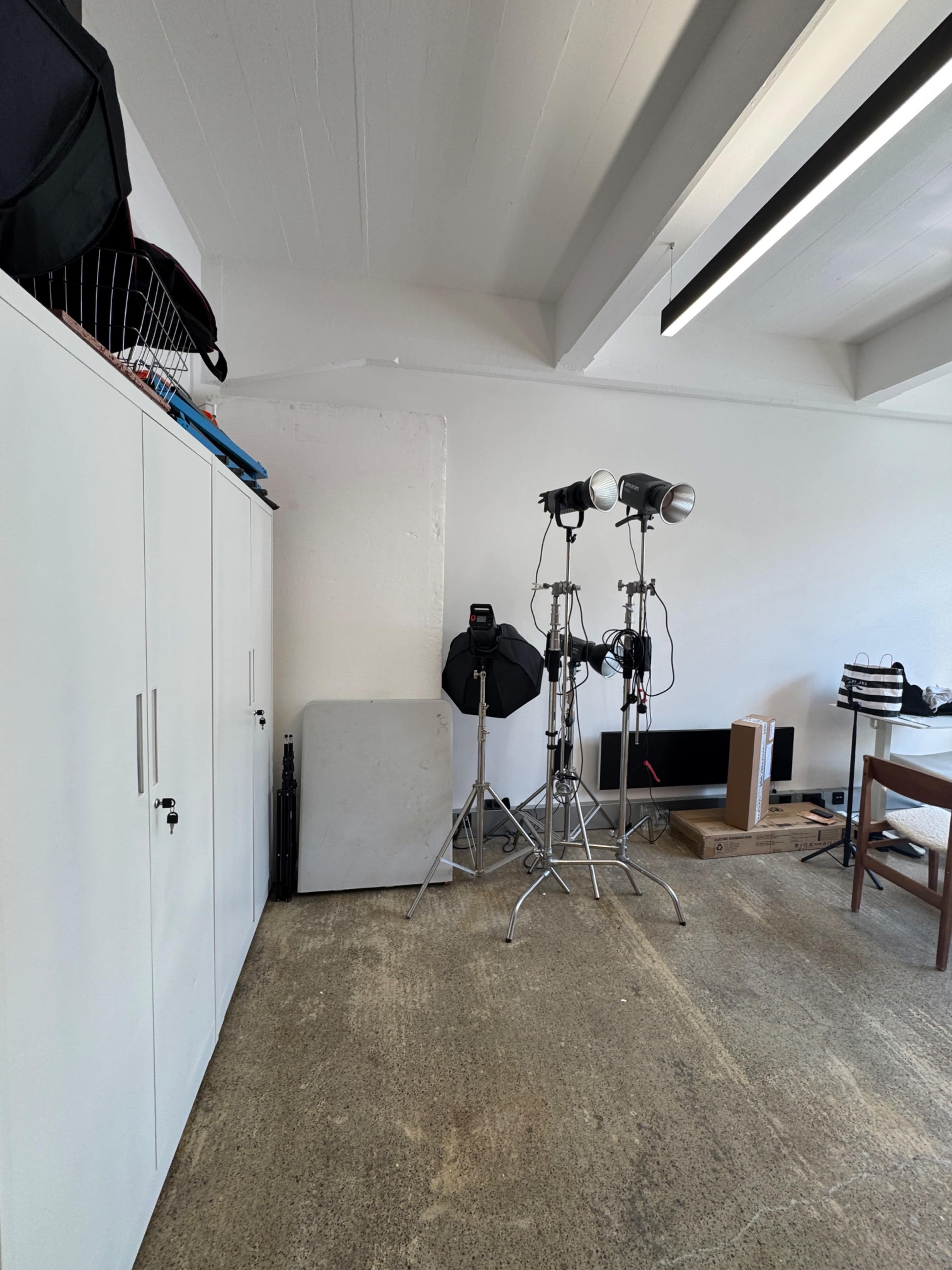The image shows a well-lit room with a white storage cabinet, three studio lights on stands, and various equipment and furniture scattered around.