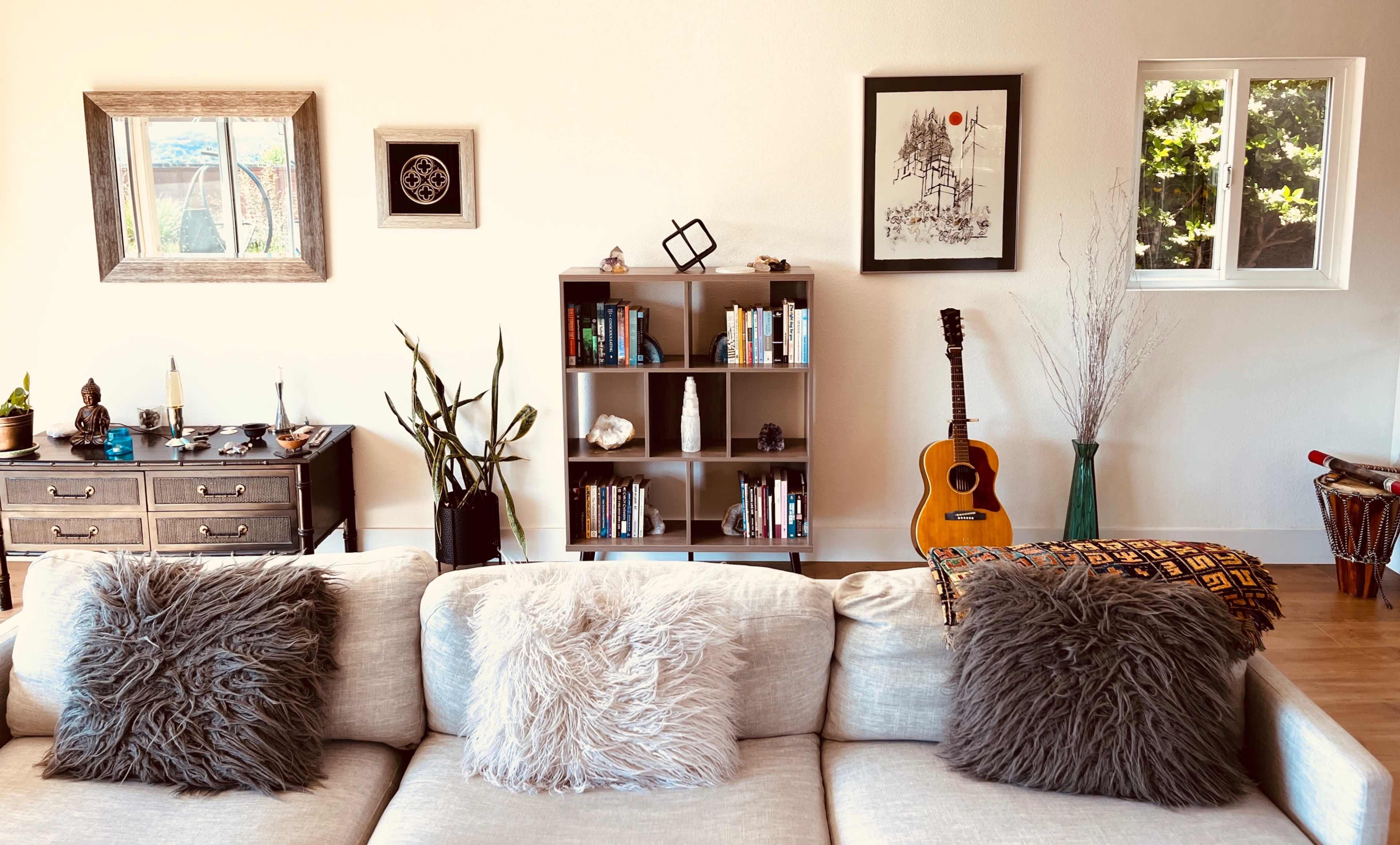 A cozy living room with a beige couch adorned with two fluffy pillows, a bookshelf filled with books, and various decorative items, including a guitar and a nearby potted plant.