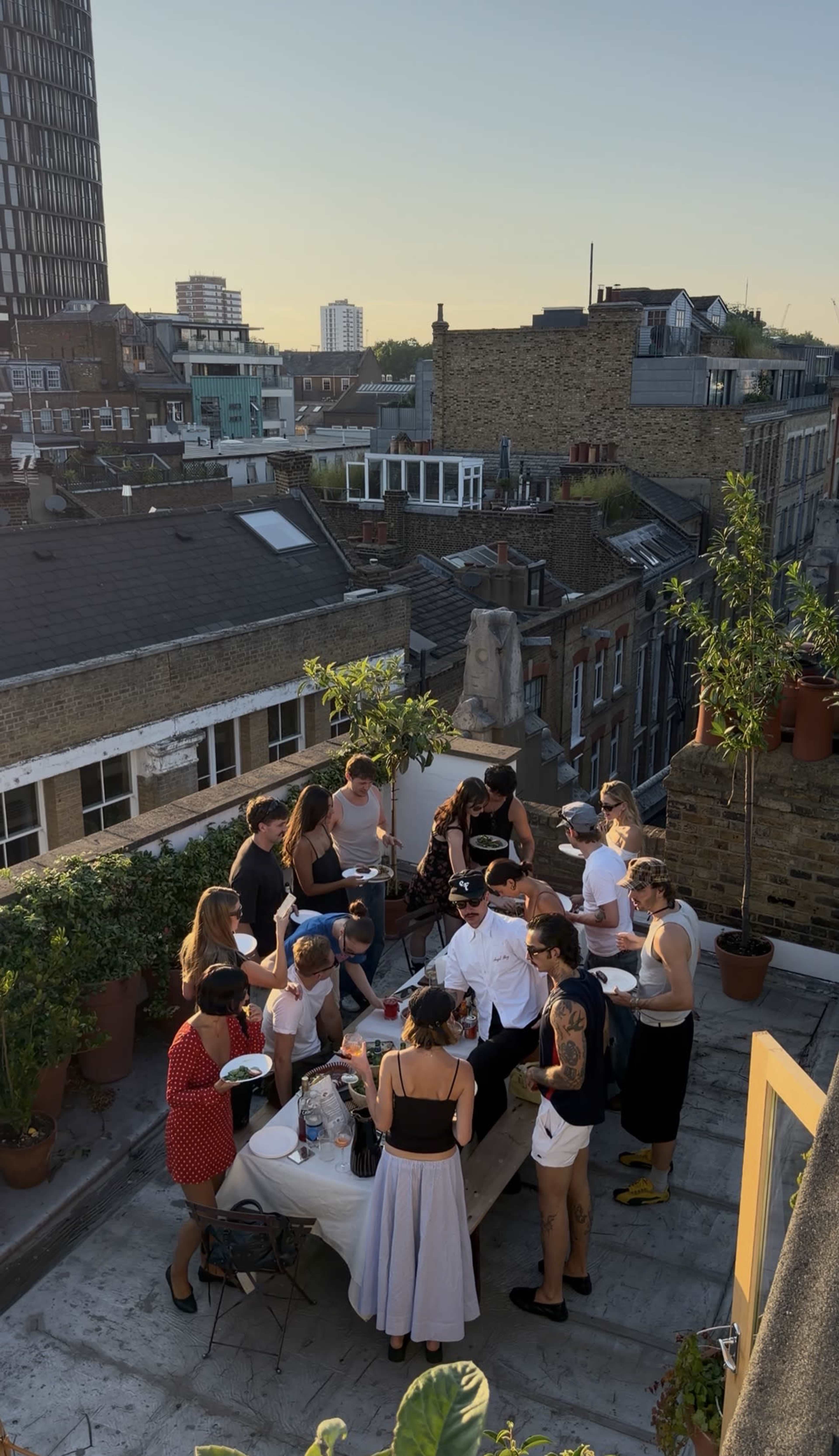 A group of people is gathered around a table on a rooftop, socializing and sharing food, with a view of city buildings in the background.