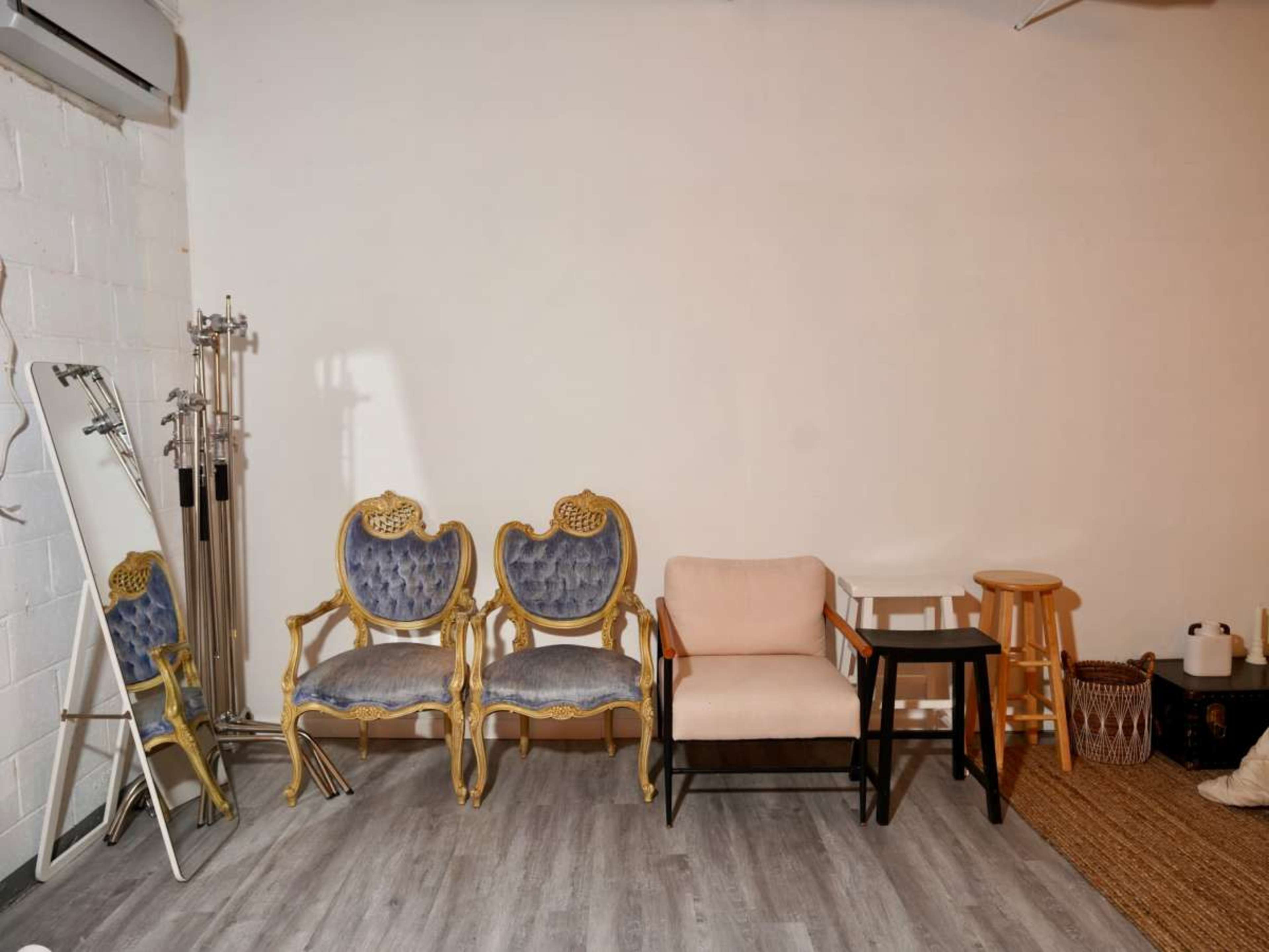 The image shows a minimalist interior space with two ornate chairs, a simple armchair, a stool, a mirror, and a woven rug.