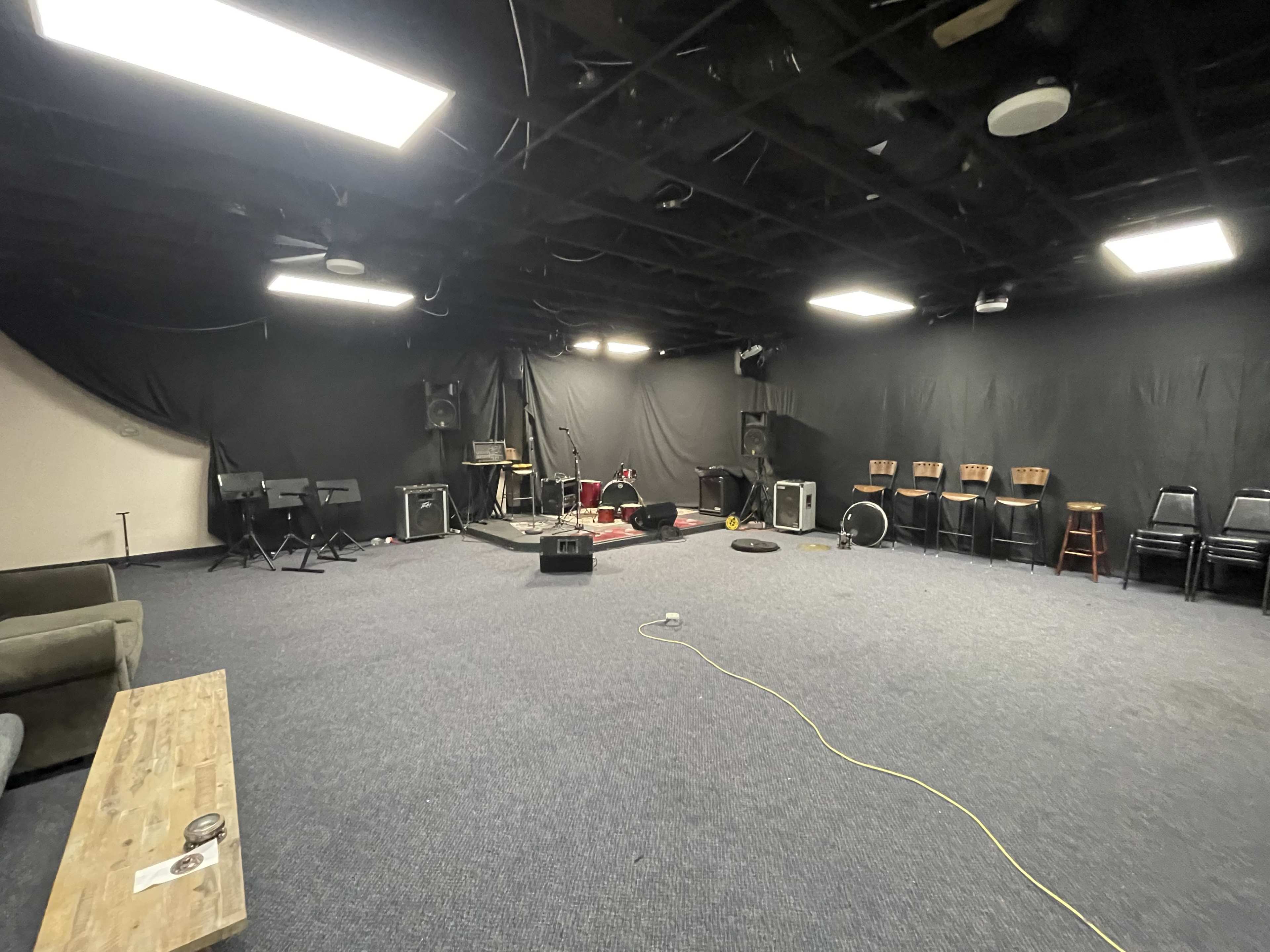The image shows a large, empty rehearsal space with black walls, several musical instruments set up in the center, and seating along the perimeter.