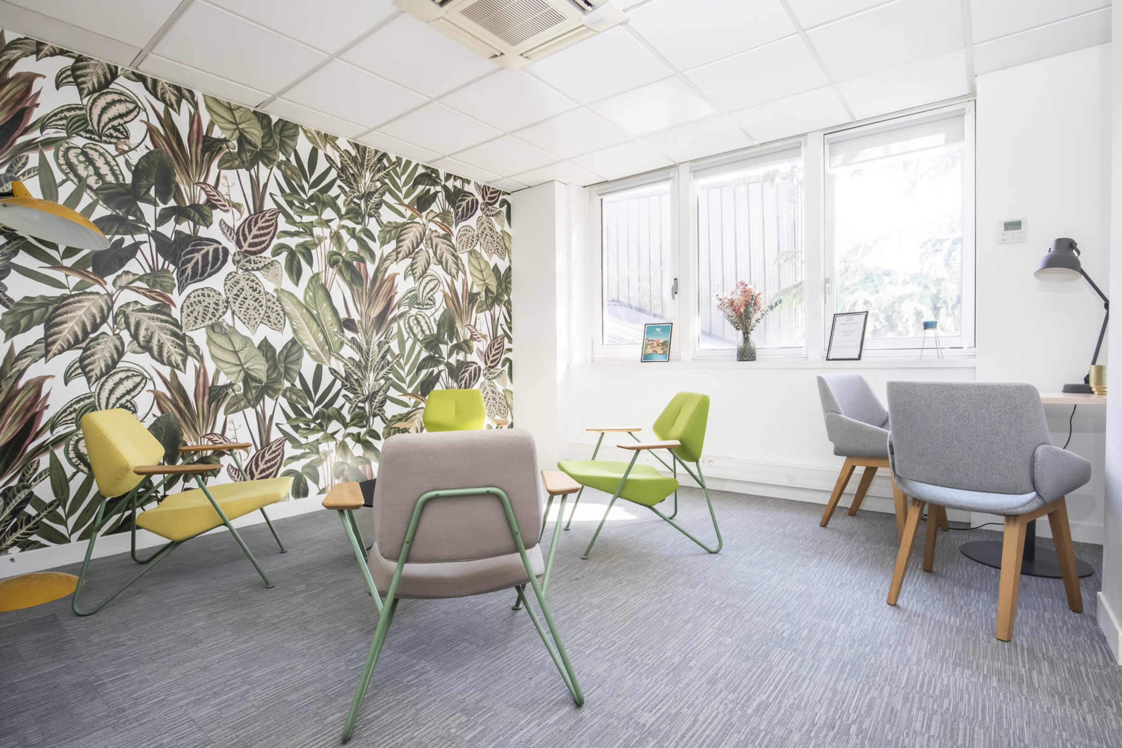 A modern office space with a tropical leaf-patterned wall, featuring several chairs in various colors arranged around a wooden table and a window with natural light.
