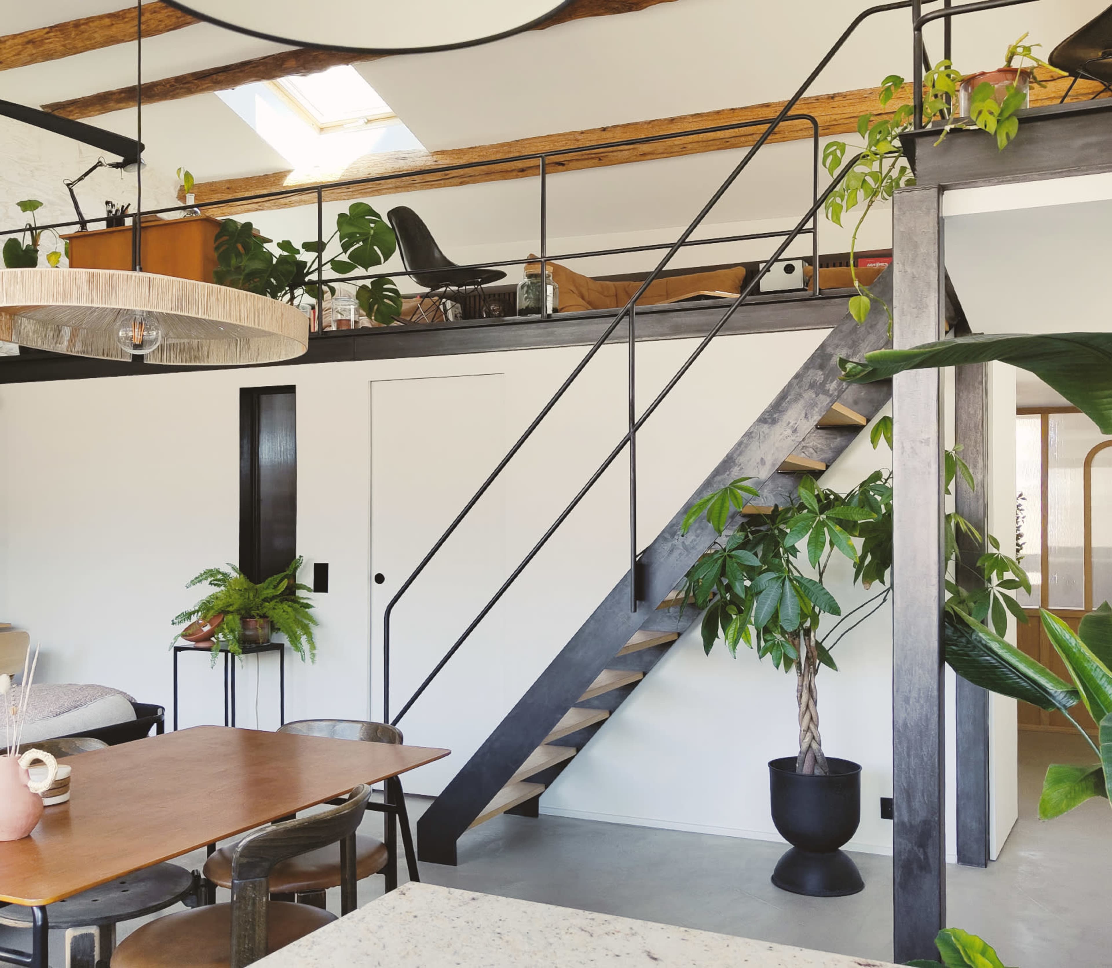 The image shows a modern interior with a staircase leading to an upper level, surrounded by plants and light-colored furnishings.