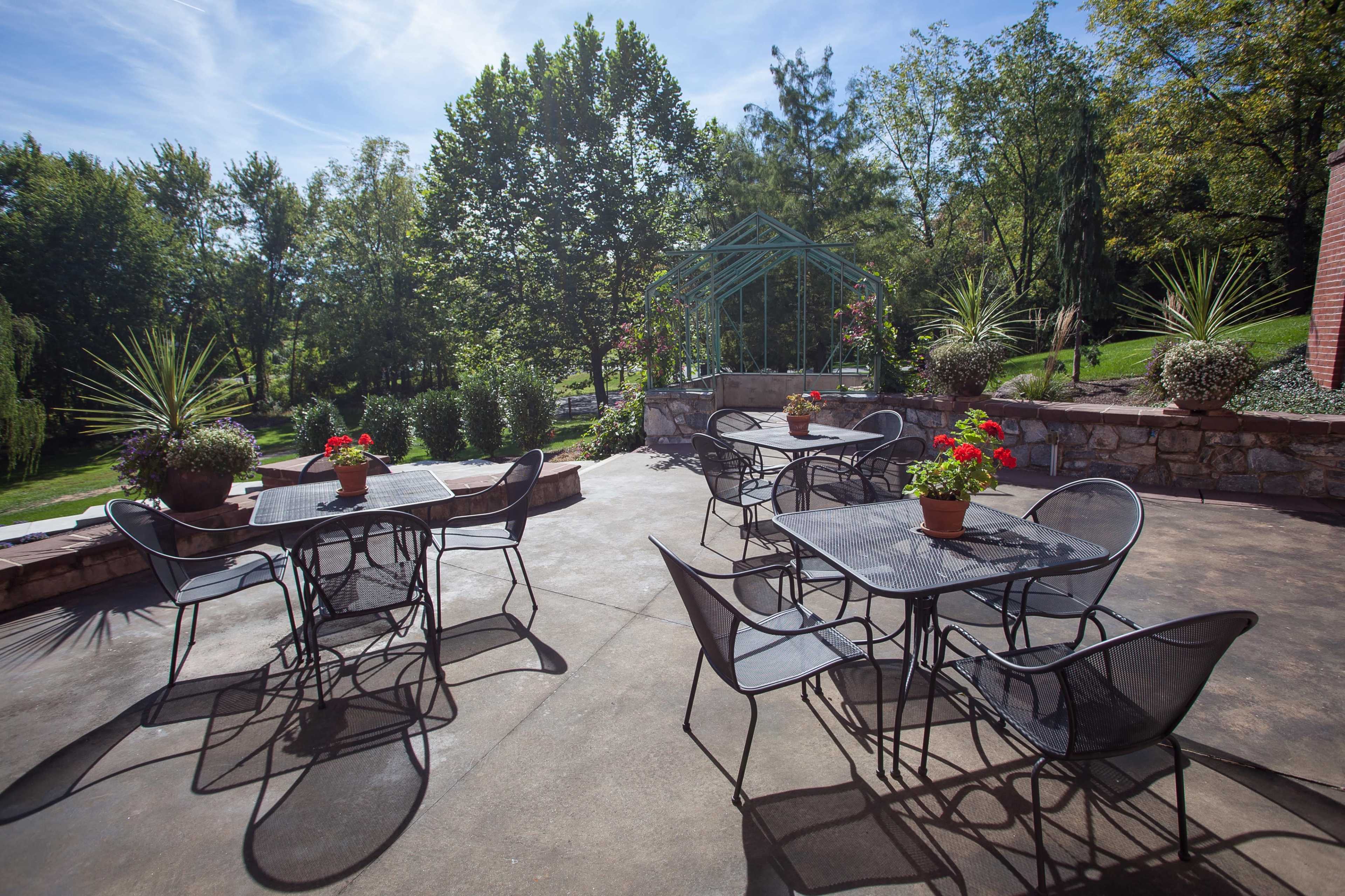 A patio area features several metal tables and chairs arranged with potted plants, surrounded by greenery and trees.