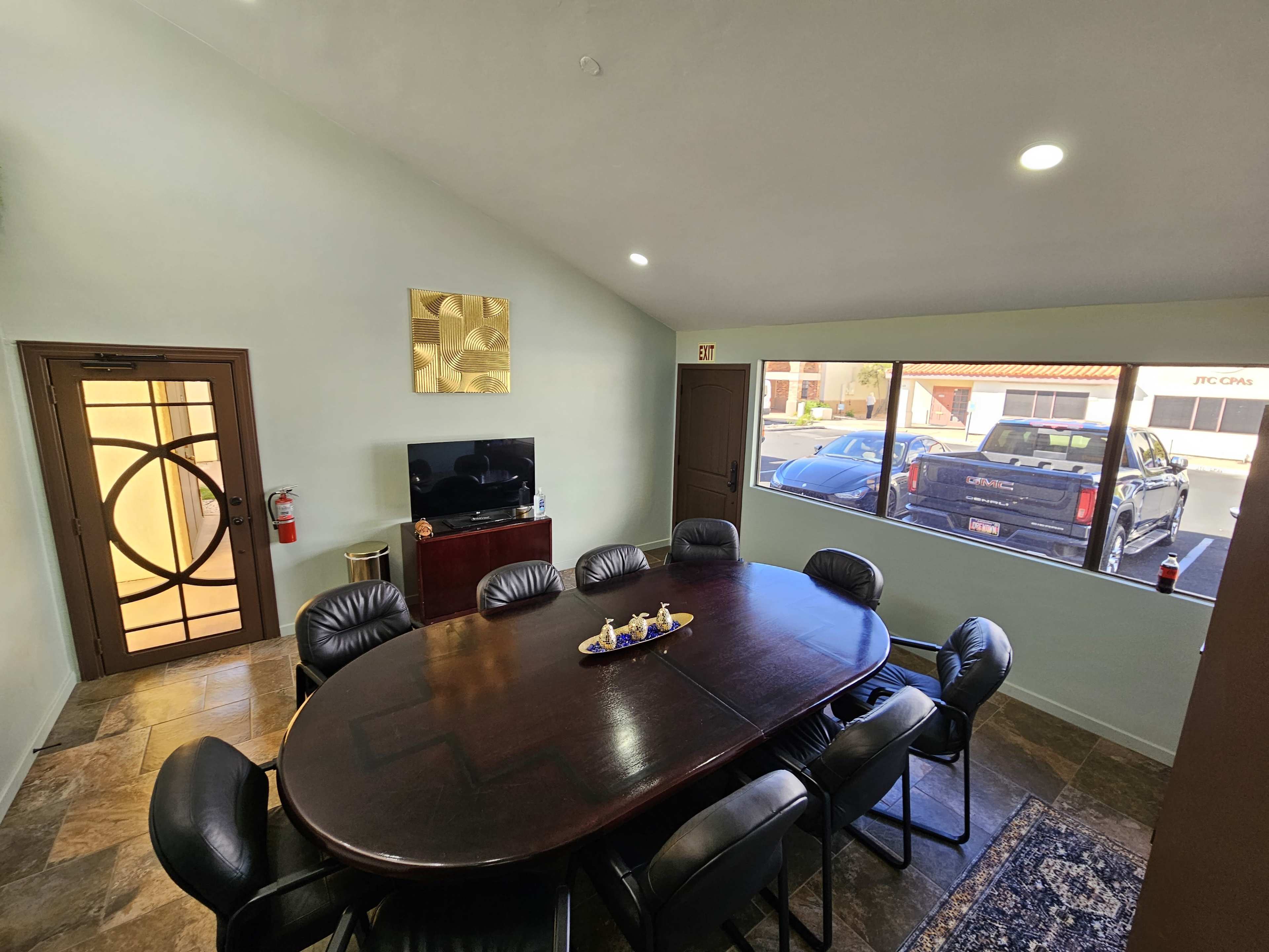 The image shows a conference room with a large oval table surrounded by black leather chairs, a television mounted on the wall, and a view of parked vehicles outside through a large window.