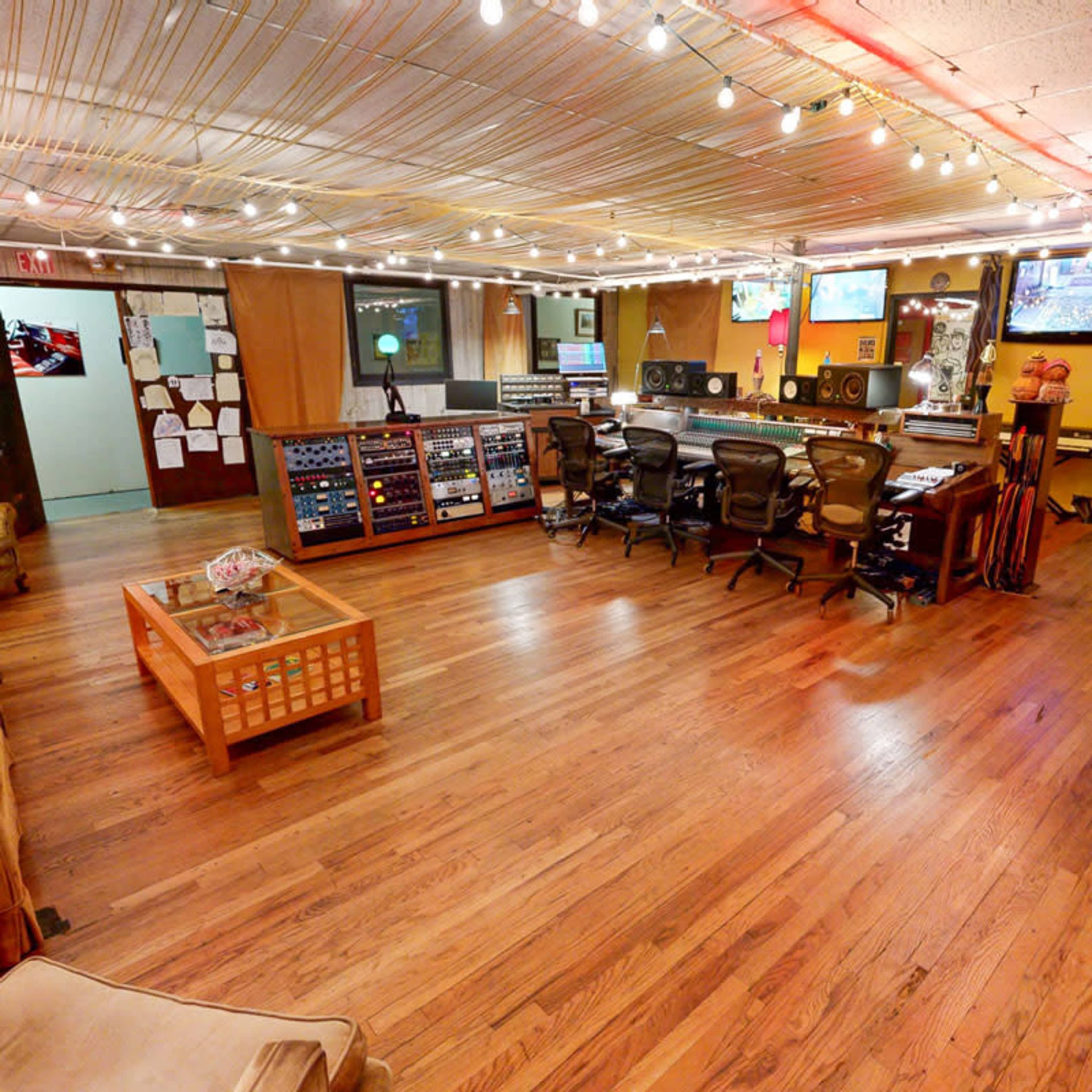 The image shows a music recording studio with a wooden floor, various audio equipment, and a seating area.