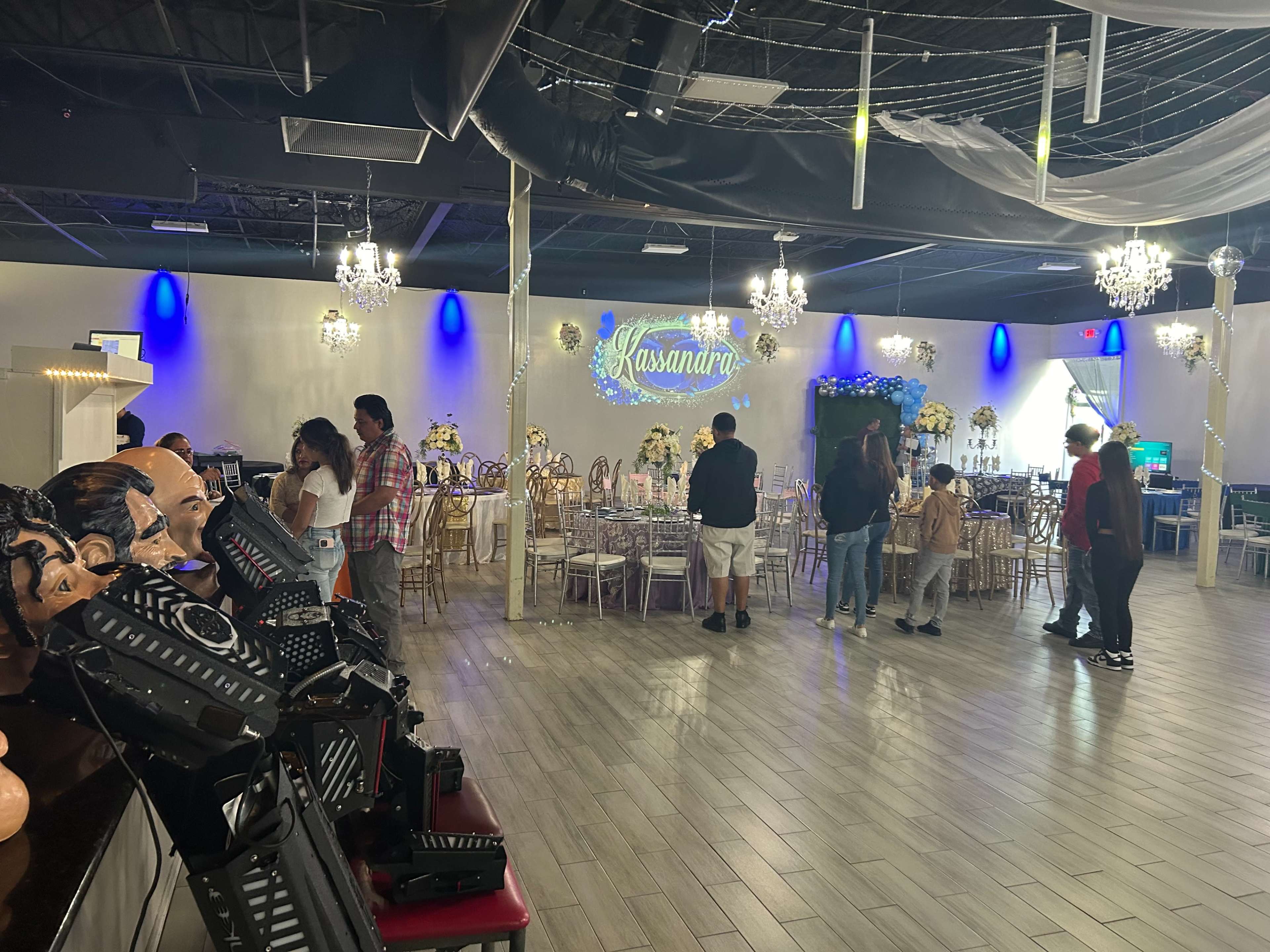 A decorated event space with tables set for a celebration and guests mingling, while a banner reading "Kassandra" is illuminated on the wall.