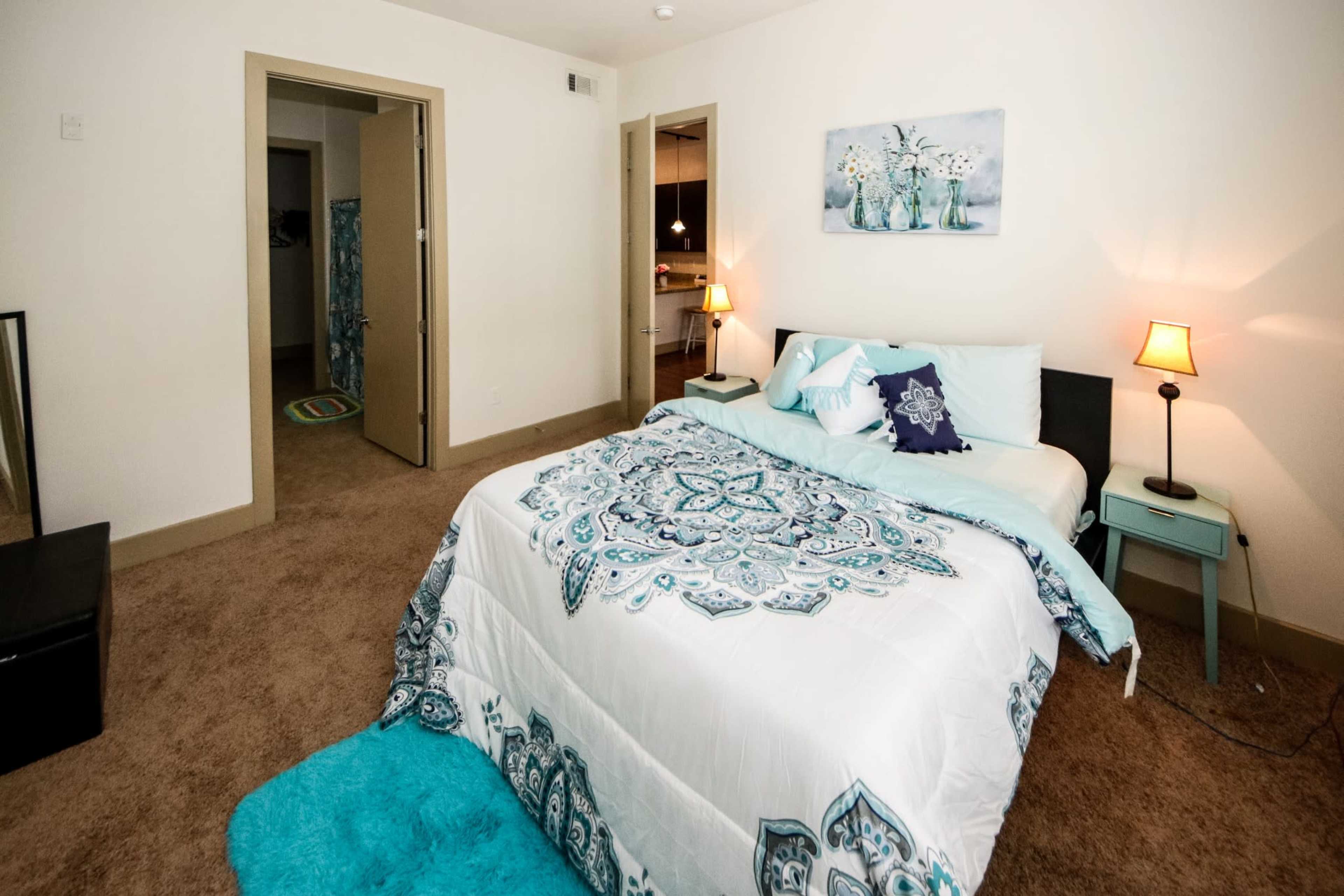 The room features a bed with a patterned spread, a nightstand with a lamp, and a doorway leading to a bathroom.