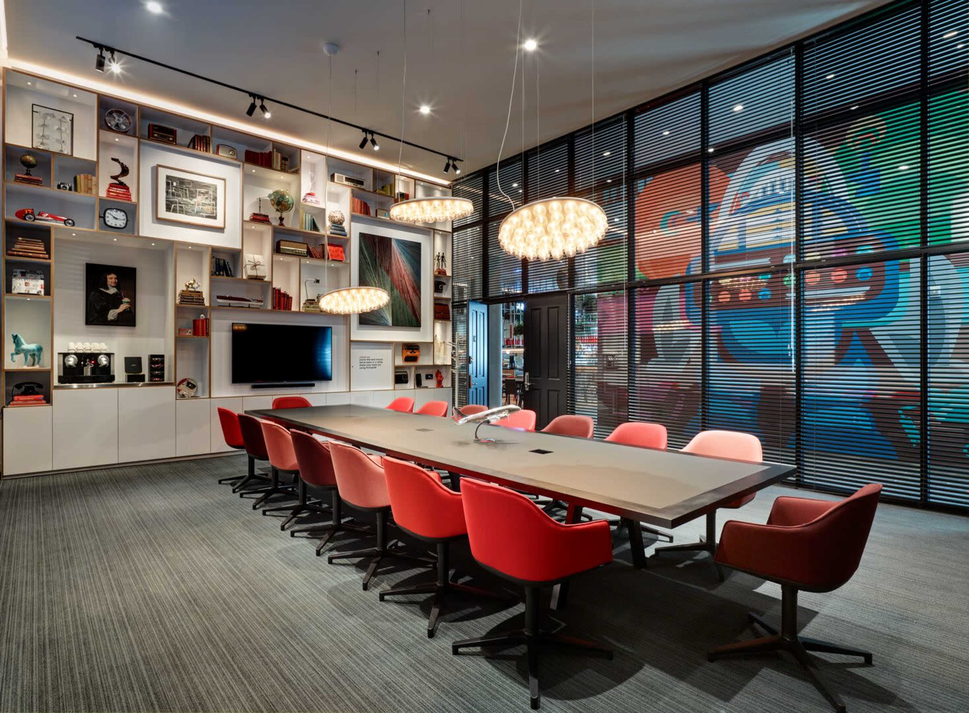 A modern conference room features a long table surrounded by red chairs, with a wall of shelves displaying art and decor, and large windows overlooking a colorful mural.