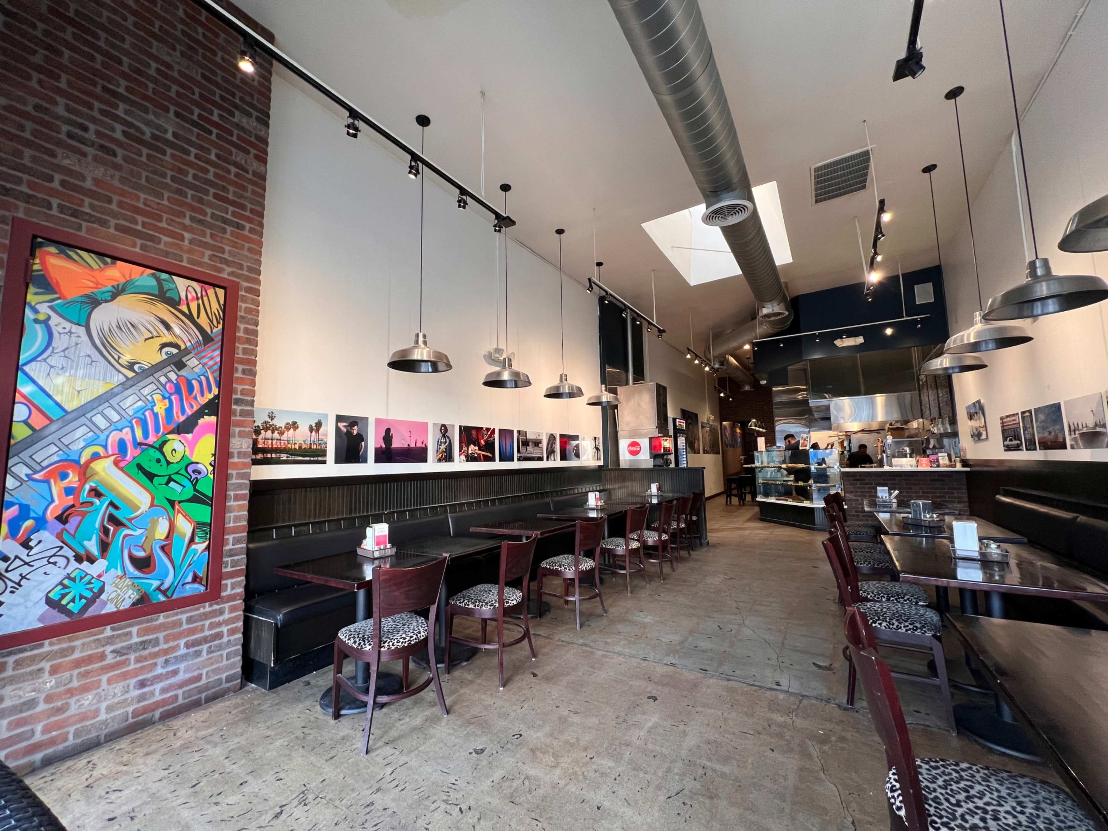 The image depicts a modern restaurant interior with exposed brick walls, a long row of tables and chairs, and a visible kitchen area in the background.