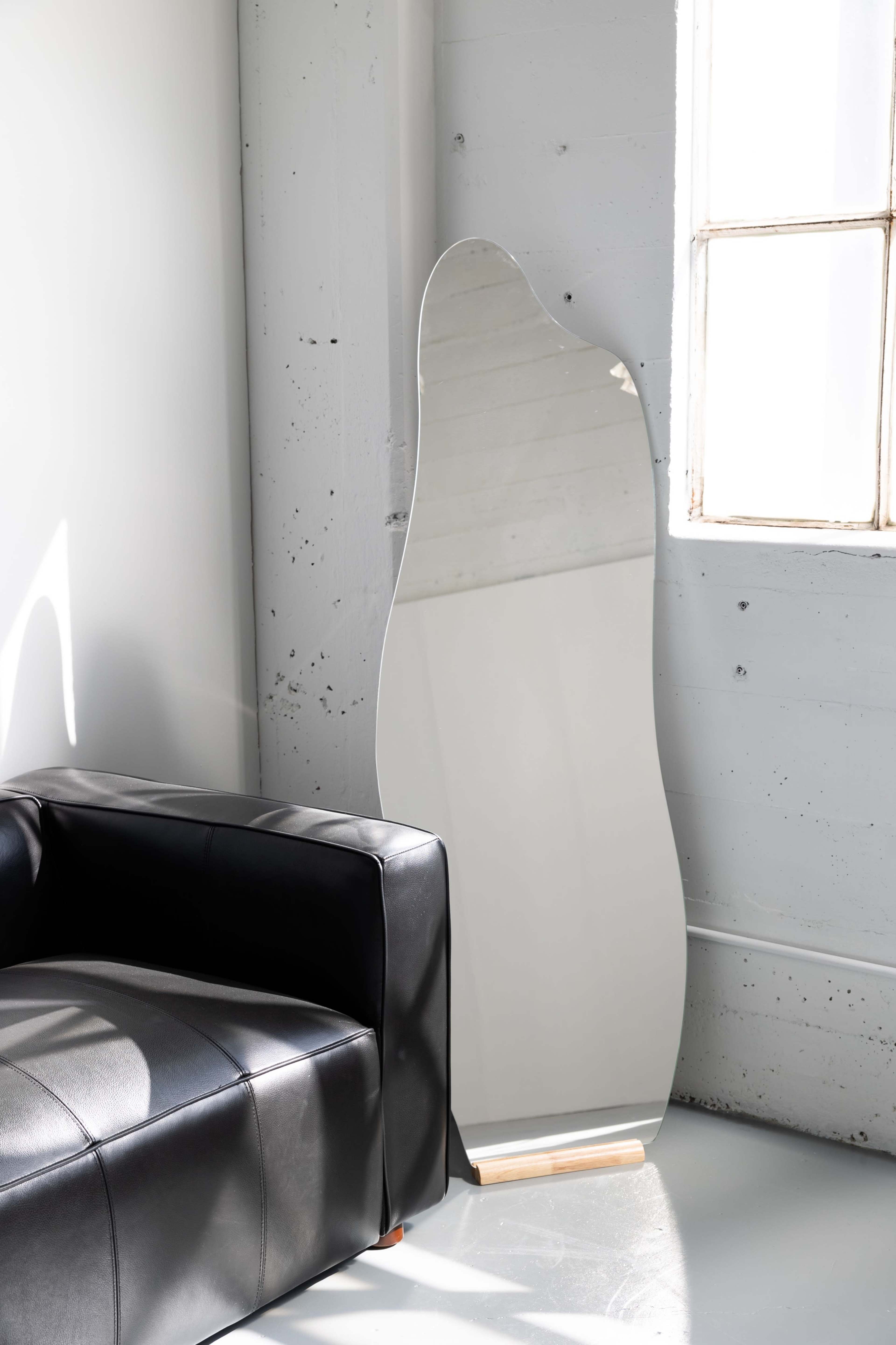A large, abstract-shaped mirror leans against a light gray wall beside a modern black leather sofa in a bright room.