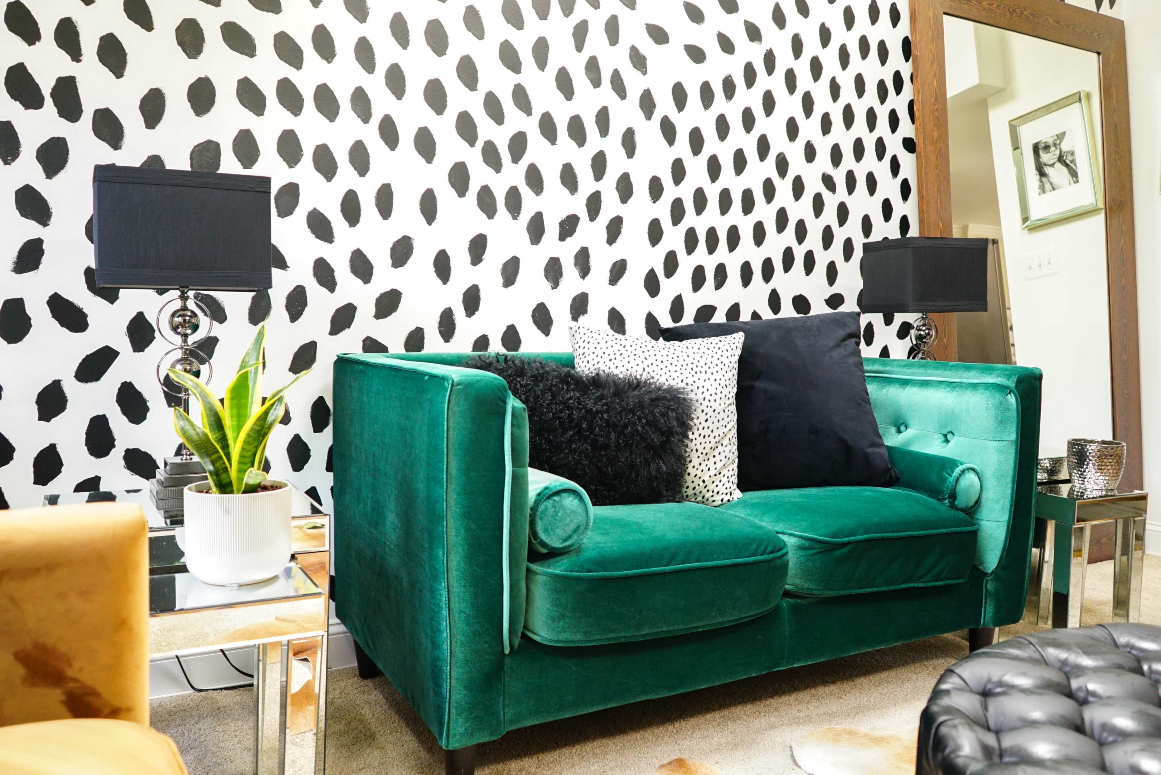 A green velvet sofa adorned with black and white pillows, set against a wall featuring a black and white spotted pattern, with two table lamps and a potted plant nearby.