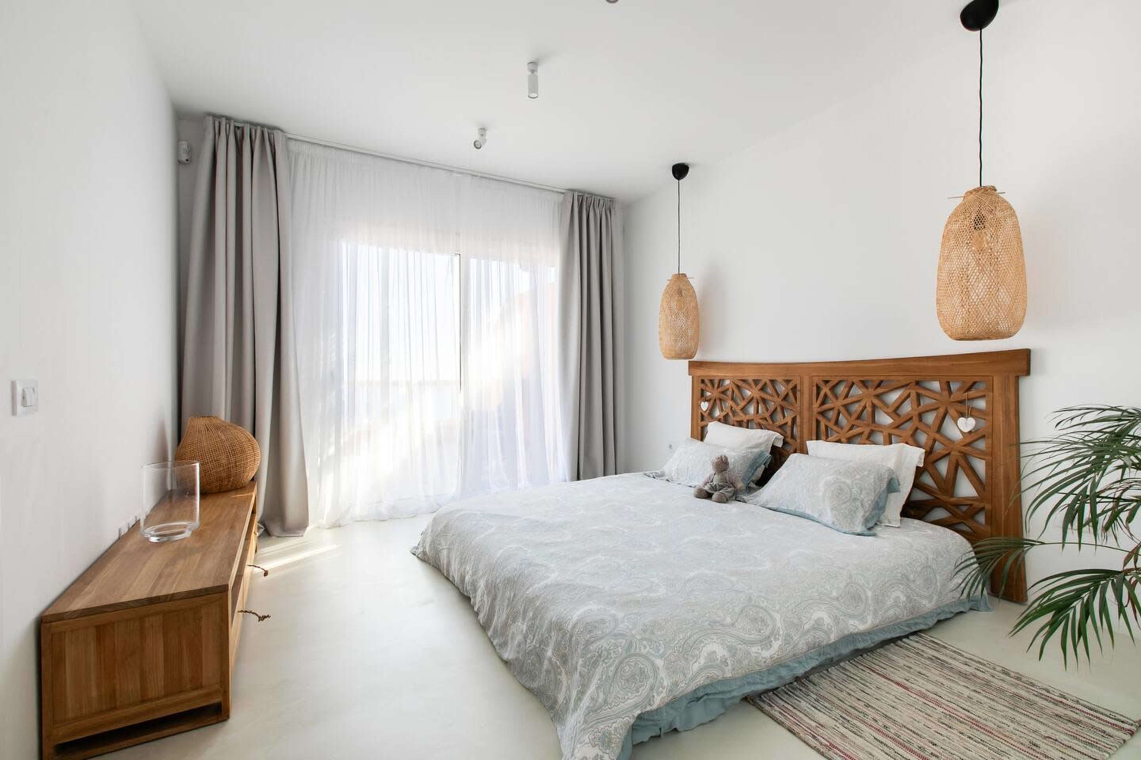 The room features a light-colored bedroom with a large bed, a wooden headboard, pendant lights, and large windows covered with sheer curtains.