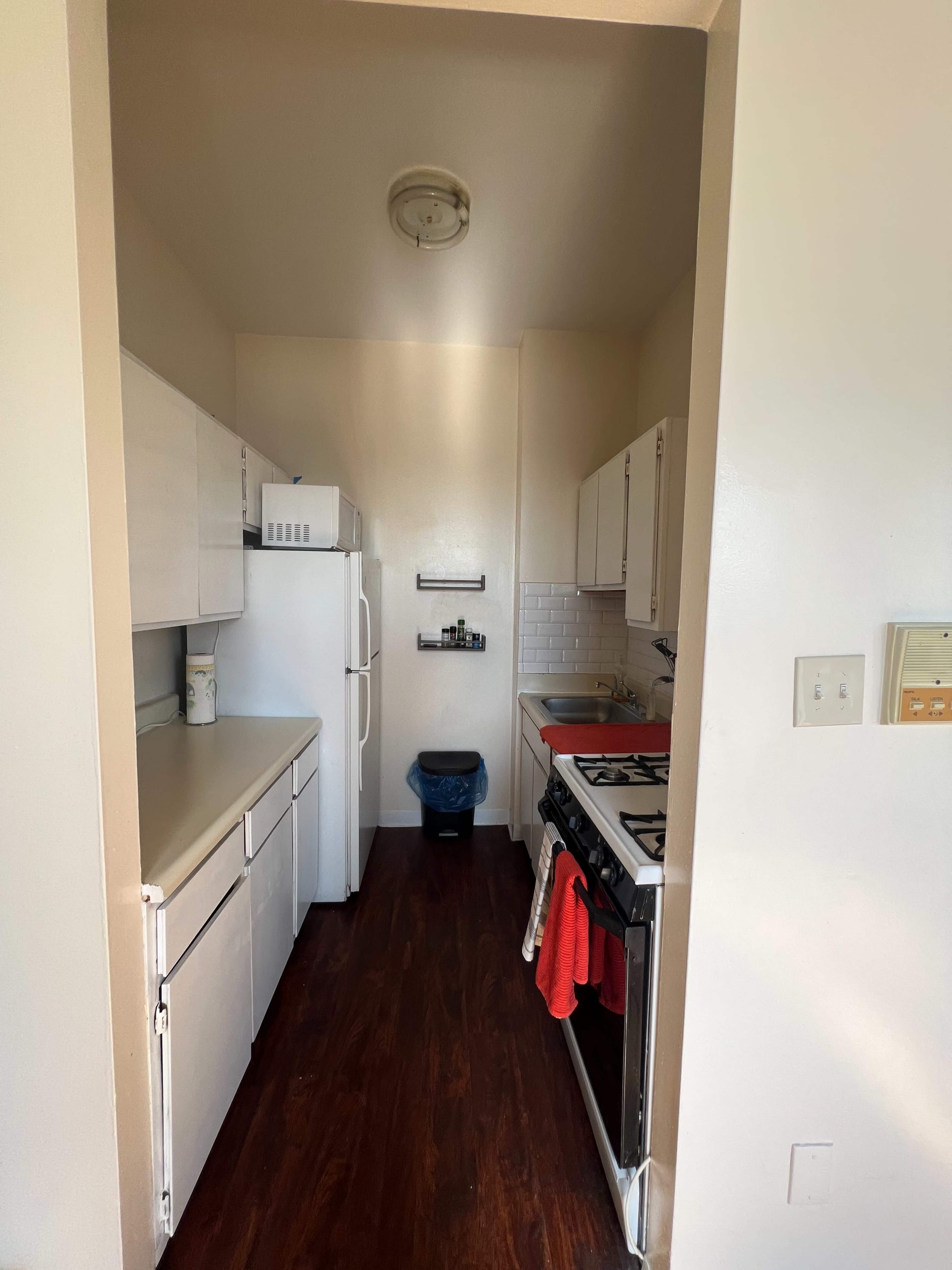 The image shows a narrow kitchen with white cabinets, an upright refrigerator, a blue trash can, a sink, and a gas stove along the wall.