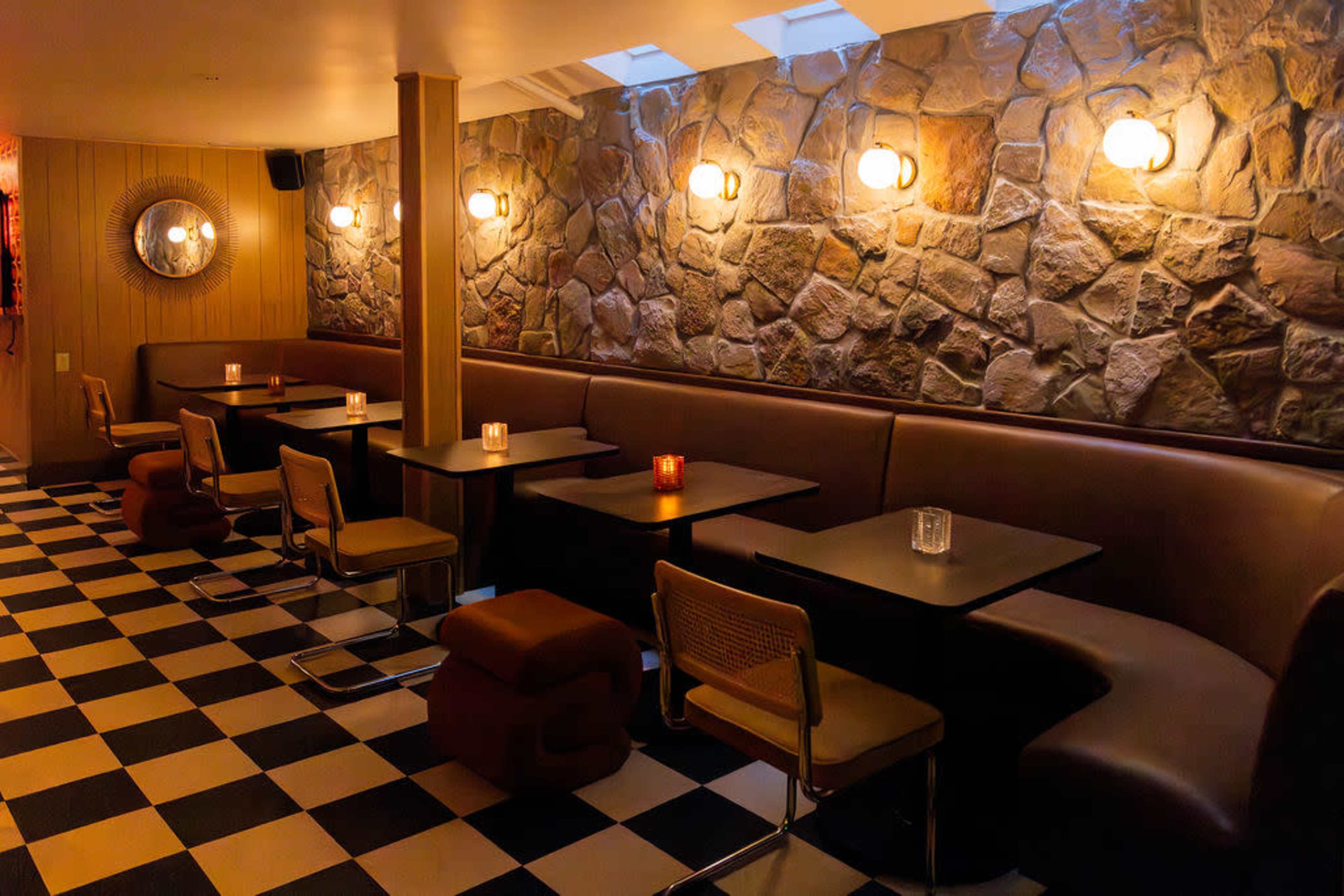 The image shows a dimly lit interior of a restaurant or cafe with stone walls, checkered flooring, and cozy seating arrangements at tables.