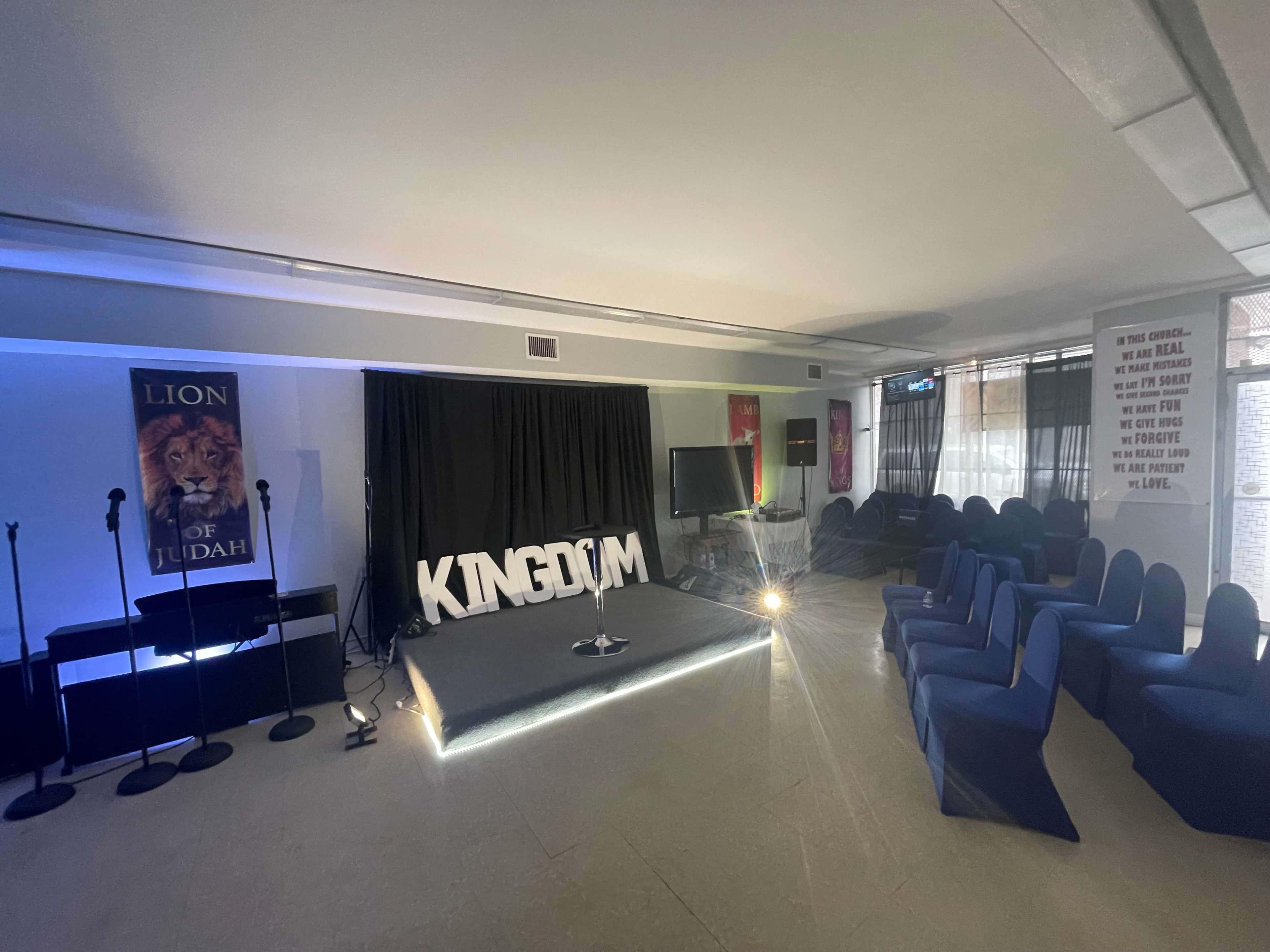 The image shows an indoor stage set up with a backdrop reading "KINGDOM," flanked by banners and microphones, in a room with blue chairs arranged for an audience.