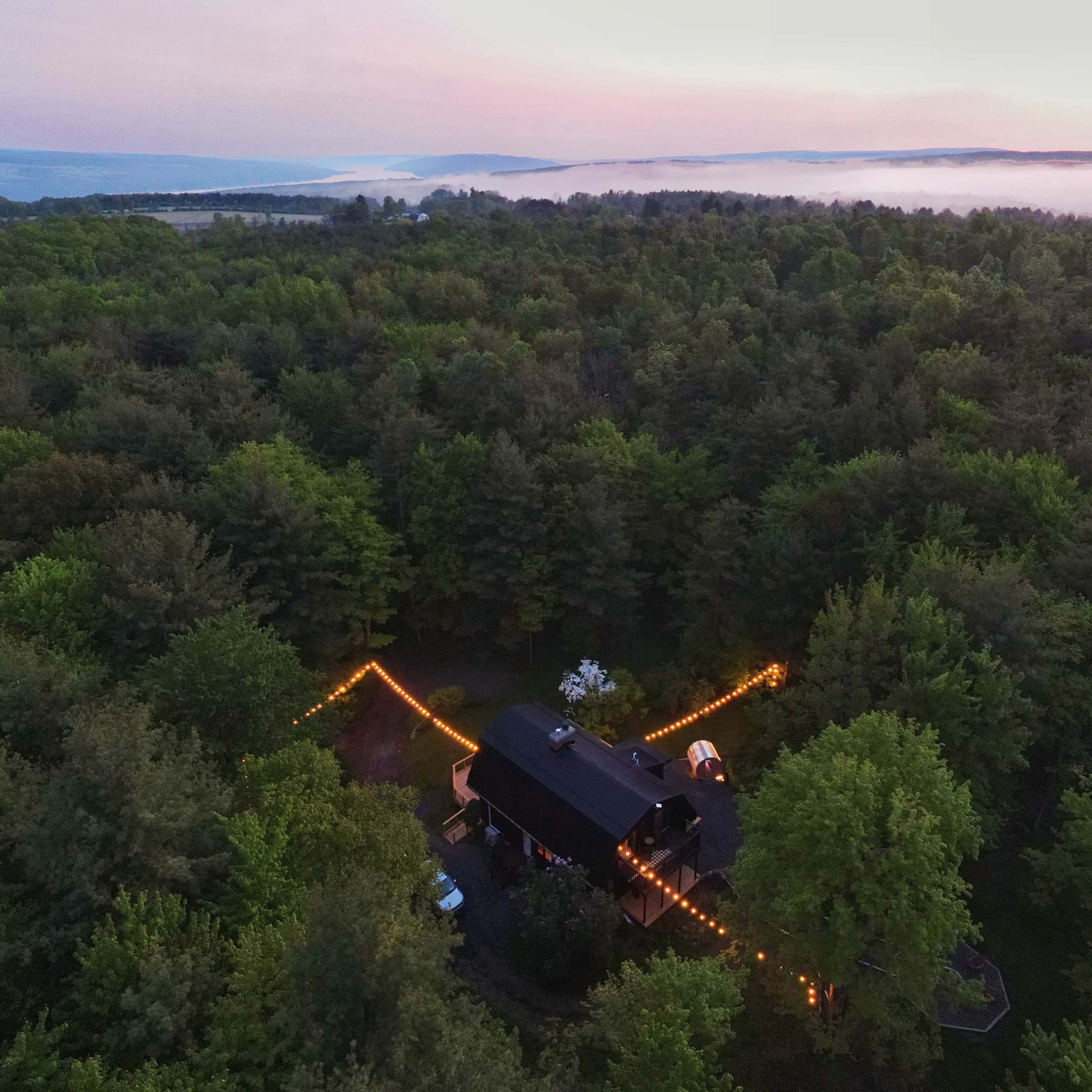 An aerial view shows a black house surrounded by dense greenery, with string lights forming a path in the yard as the sun sets over a misty landscape.