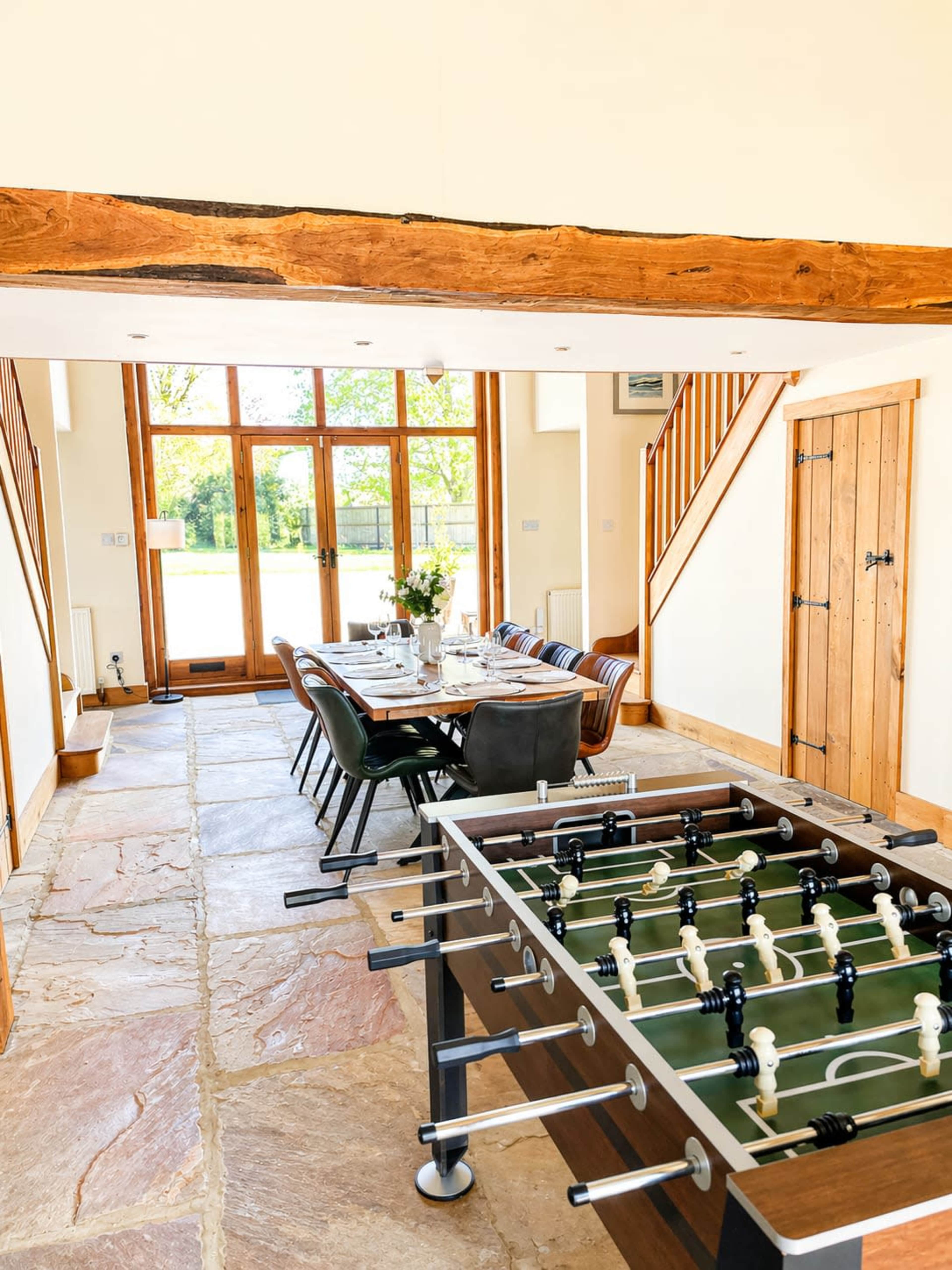 The image shows a spacious room featuring a foosball table in the foreground and a dining table set for multiple guests in the background, with large windows allowing natural light.