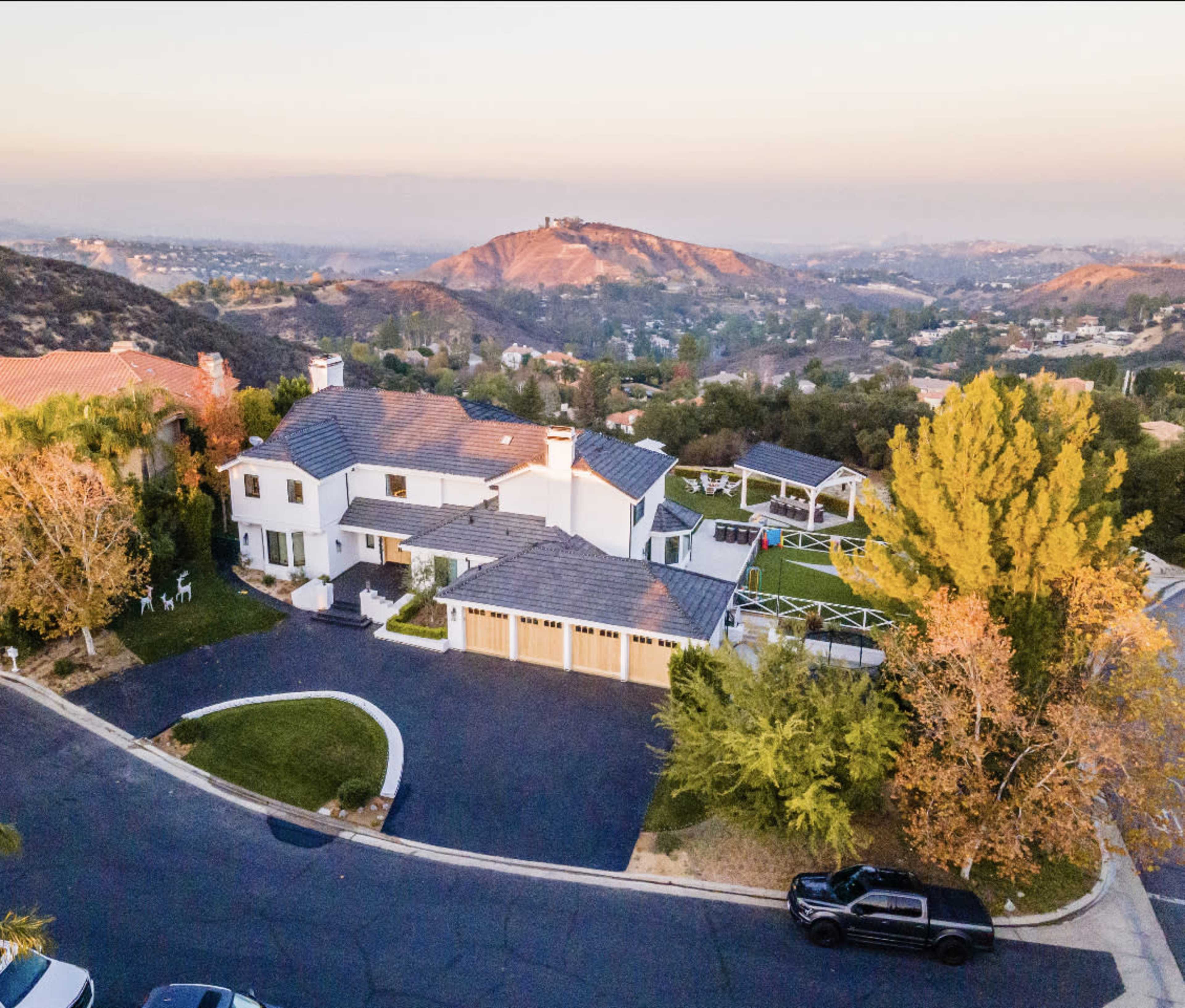 An aerial view shows a large white house with a landscaped yard, situated on a suburban street with rolling hills in the background.