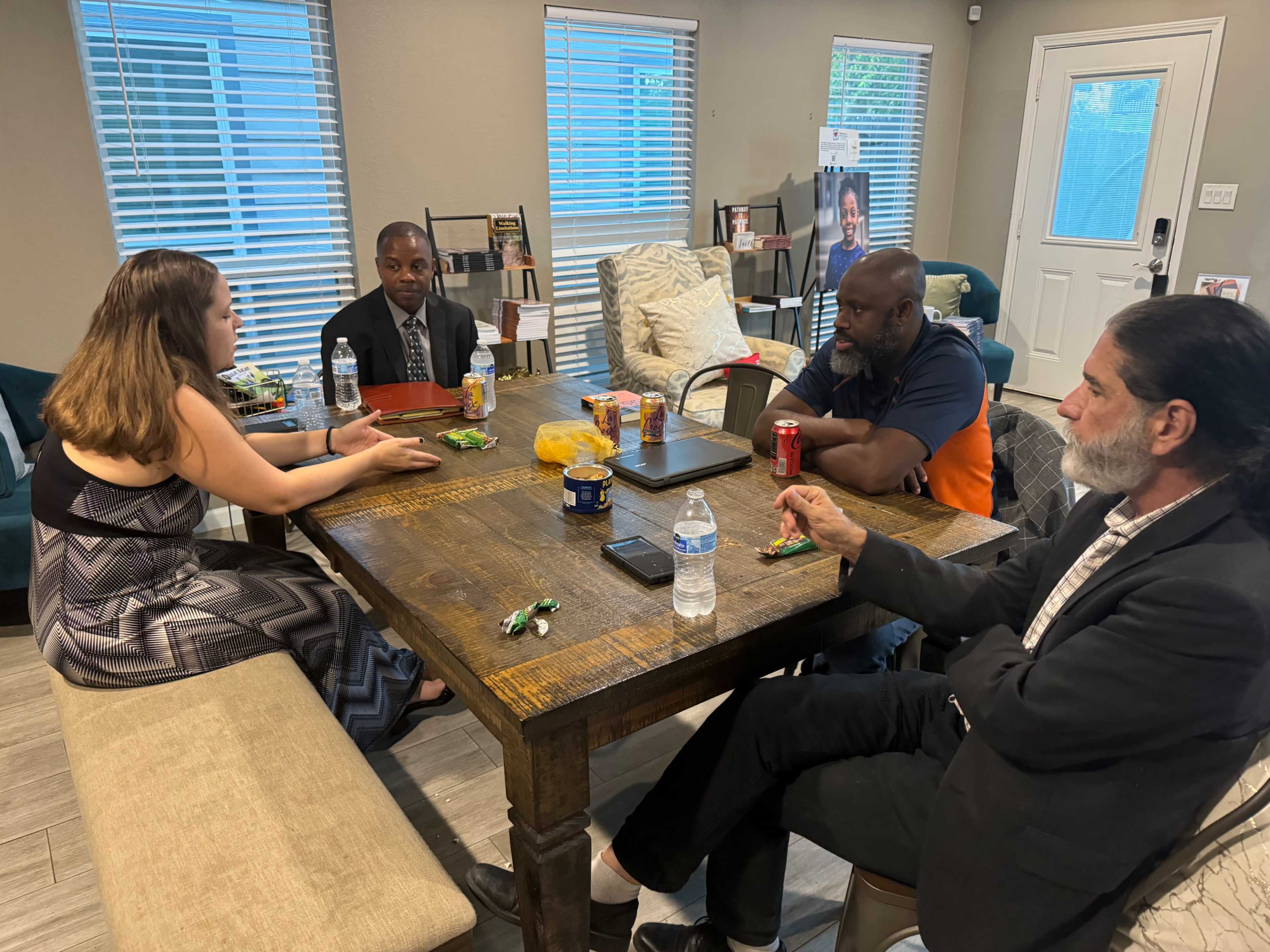 Four people are seated around a wooden table with snacks and drinks in a well-lit room.