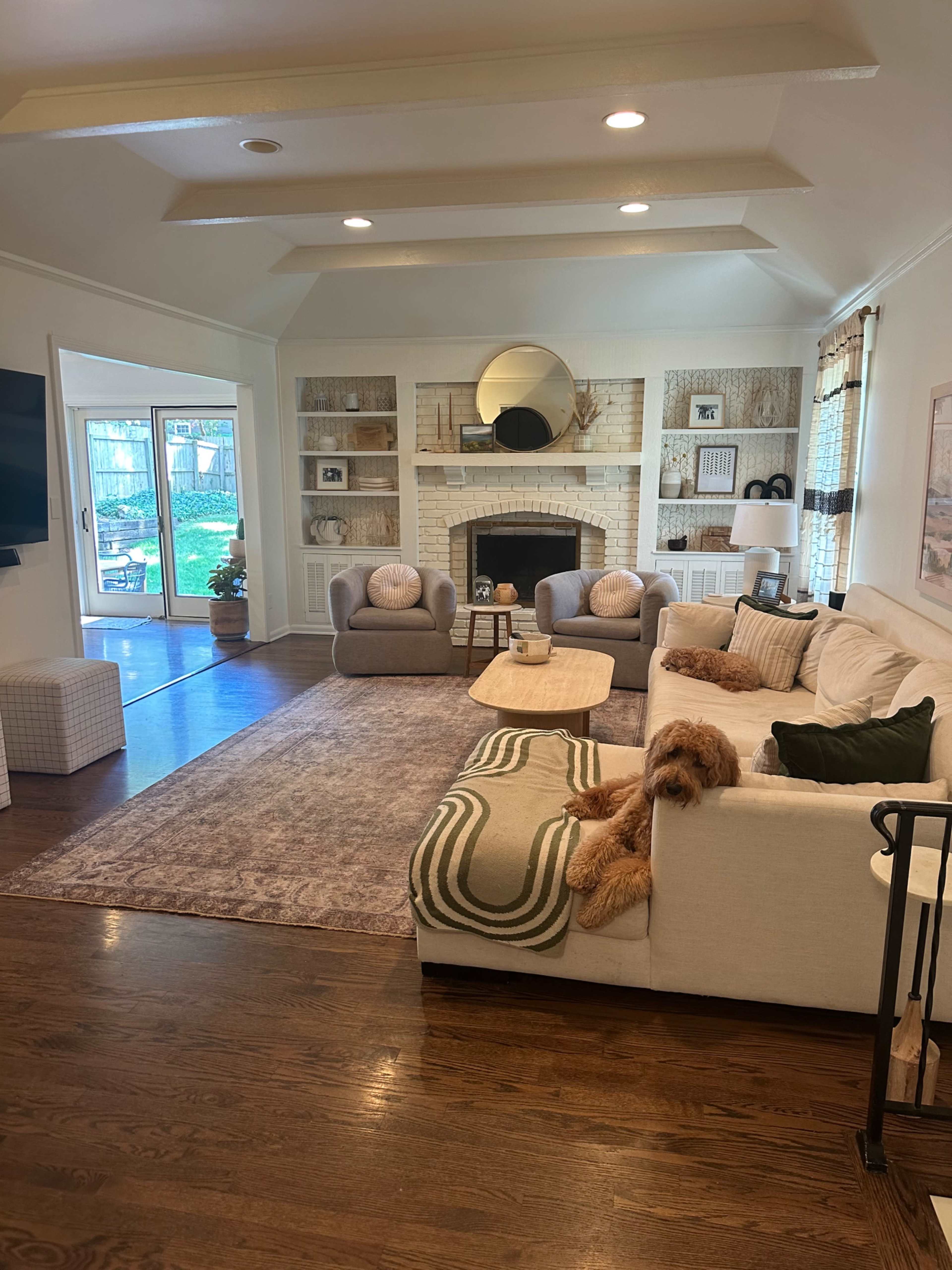 A spacious living room features a beige sofa with decorative pillows, two armchairs, a coffee table, a fireplace with a round mirror above it, and a sliding glass door leading to an outdoor area.