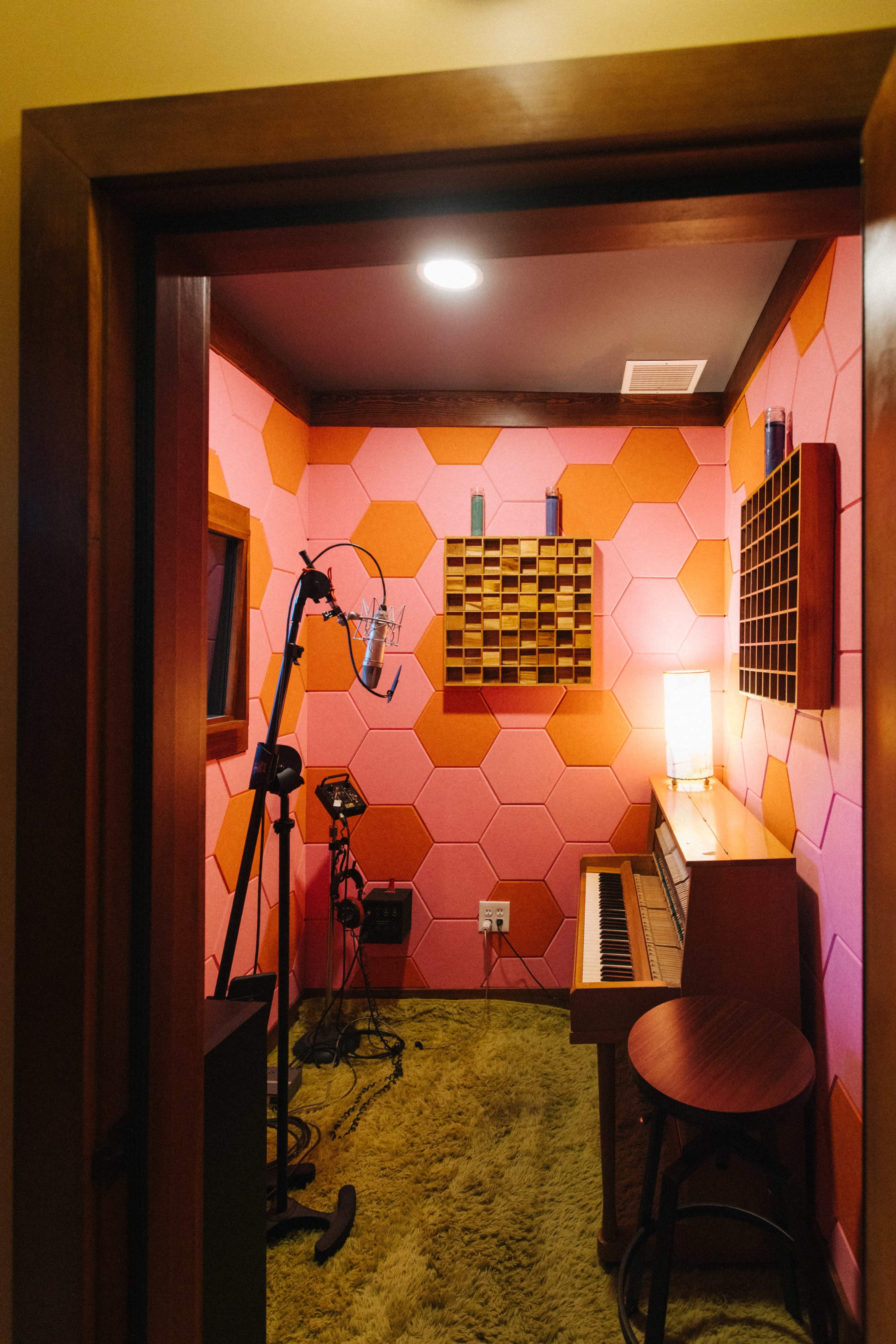 A small recording studio with pink hexagonal soundproofing panels, a microphone stand, an upright piano, and a cozy rug on the floor.