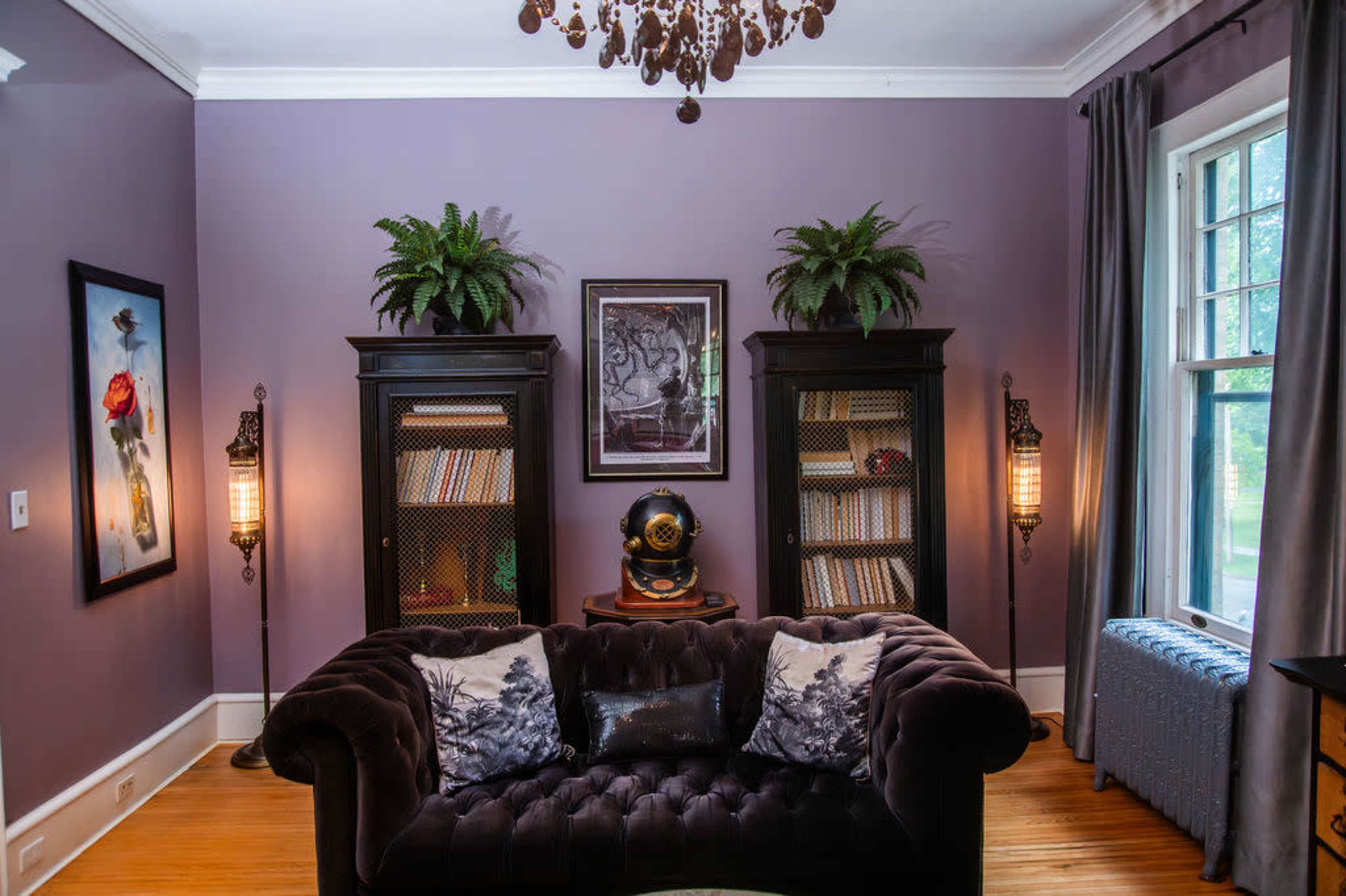 The image depicts a cozy living room with a dark velvet couch, two black bookcases filled with books, and a chandelier hanging from the ceiling, all complemented by purple walls and potted plants.