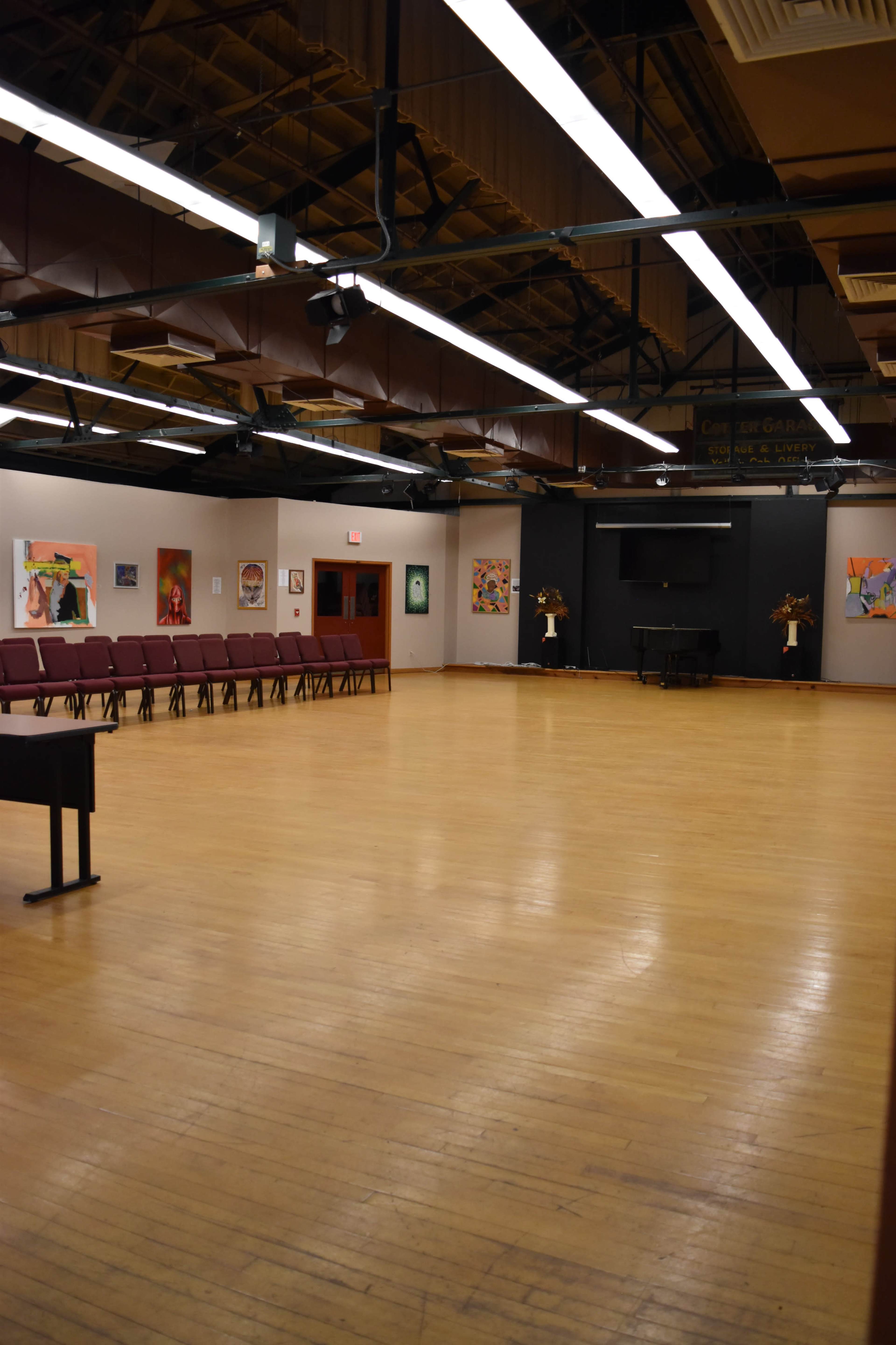 The image shows an empty hall with wooden flooring, lined with maroon chairs on one side and featuring artwork on the walls.