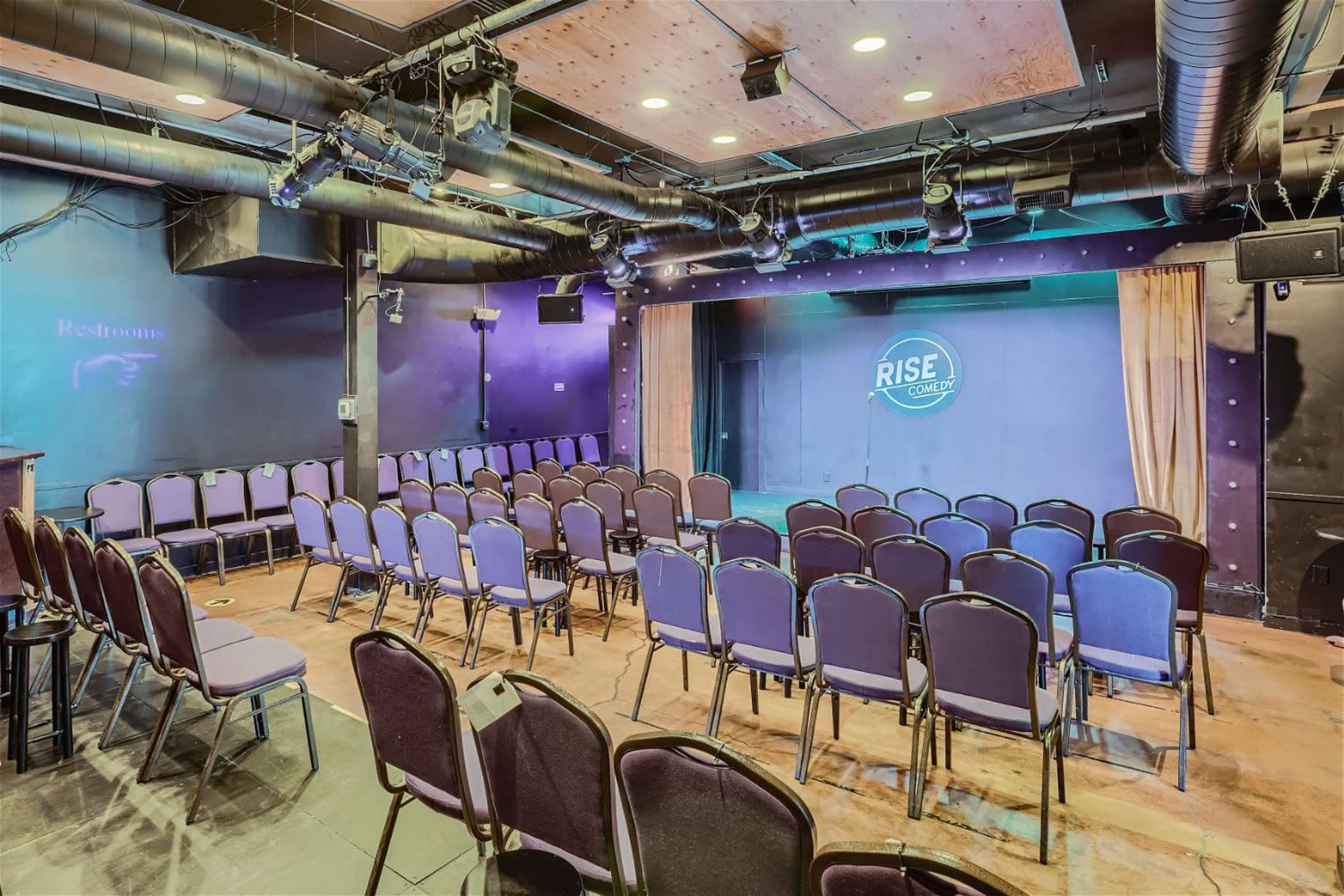 A small theater space with rows of purple chairs facing a stage, featuring a backdrop with the logo "RISE."