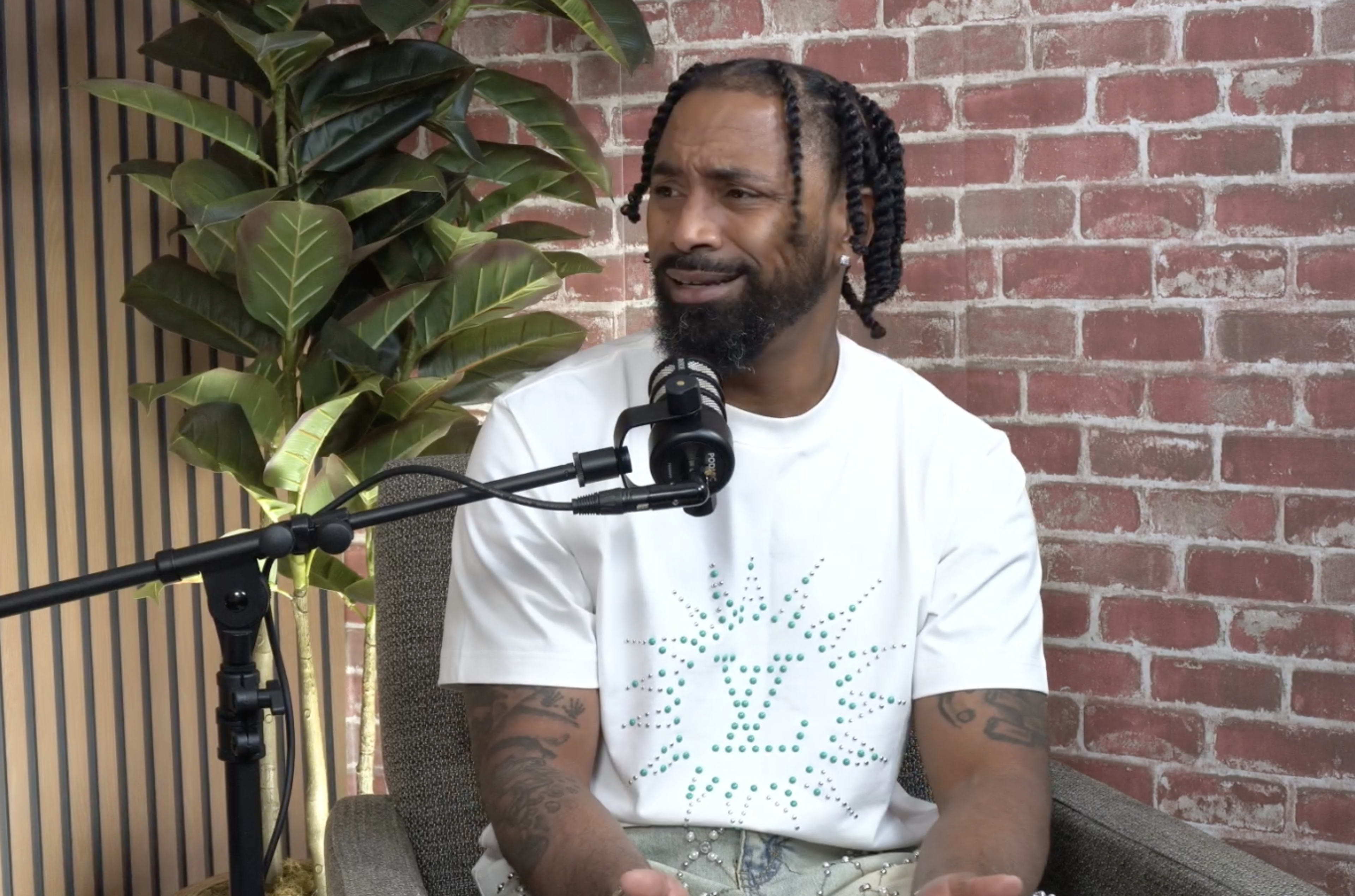 A man with braided hair sits in a chair with a microphone and a green plant nearby, gesturing as he speaks against a backdrop of brick walls.