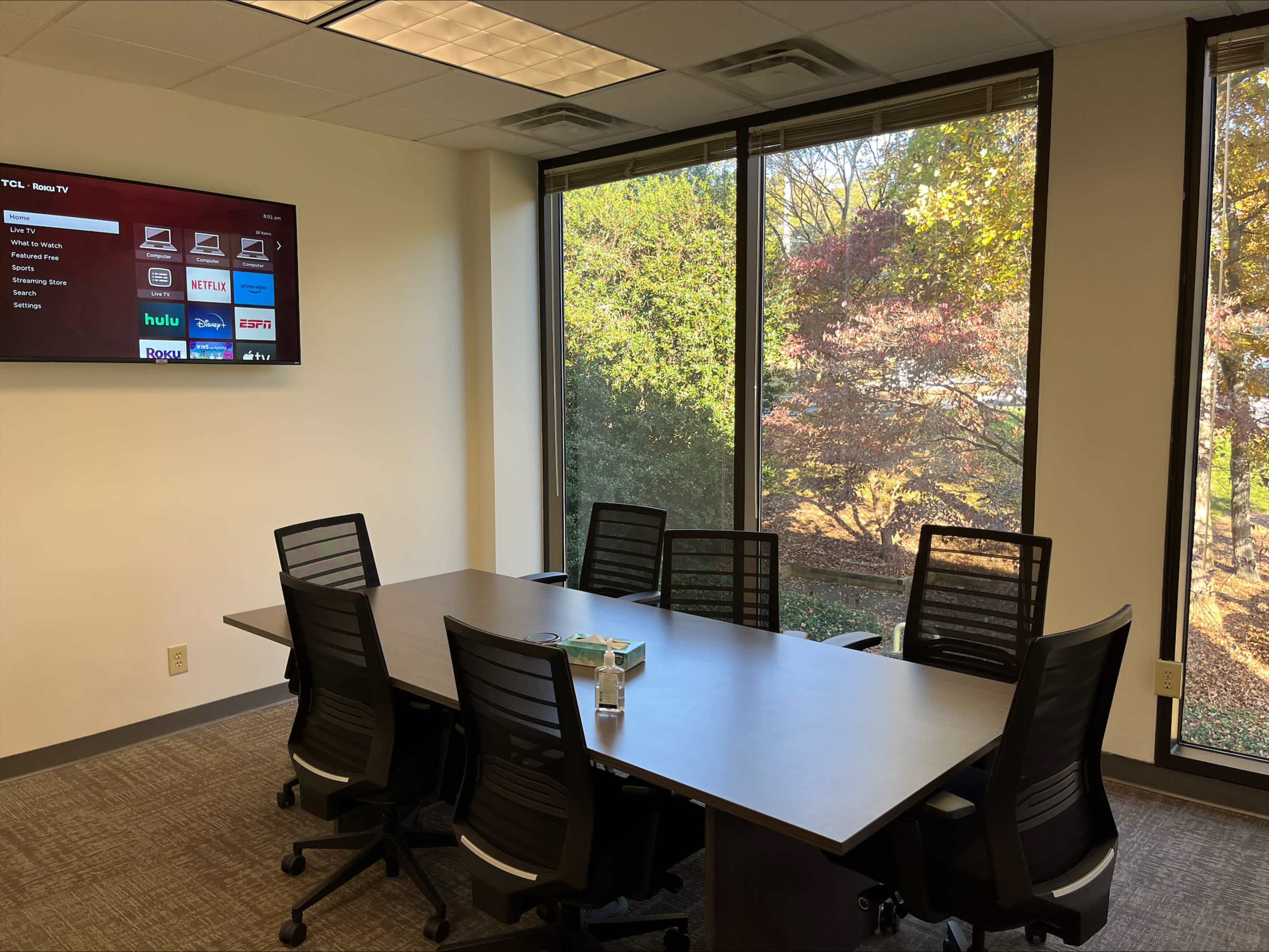 A conference room features a long table surrounded by ergonomic chairs and a wall-mounted screen displaying various streaming service logos.