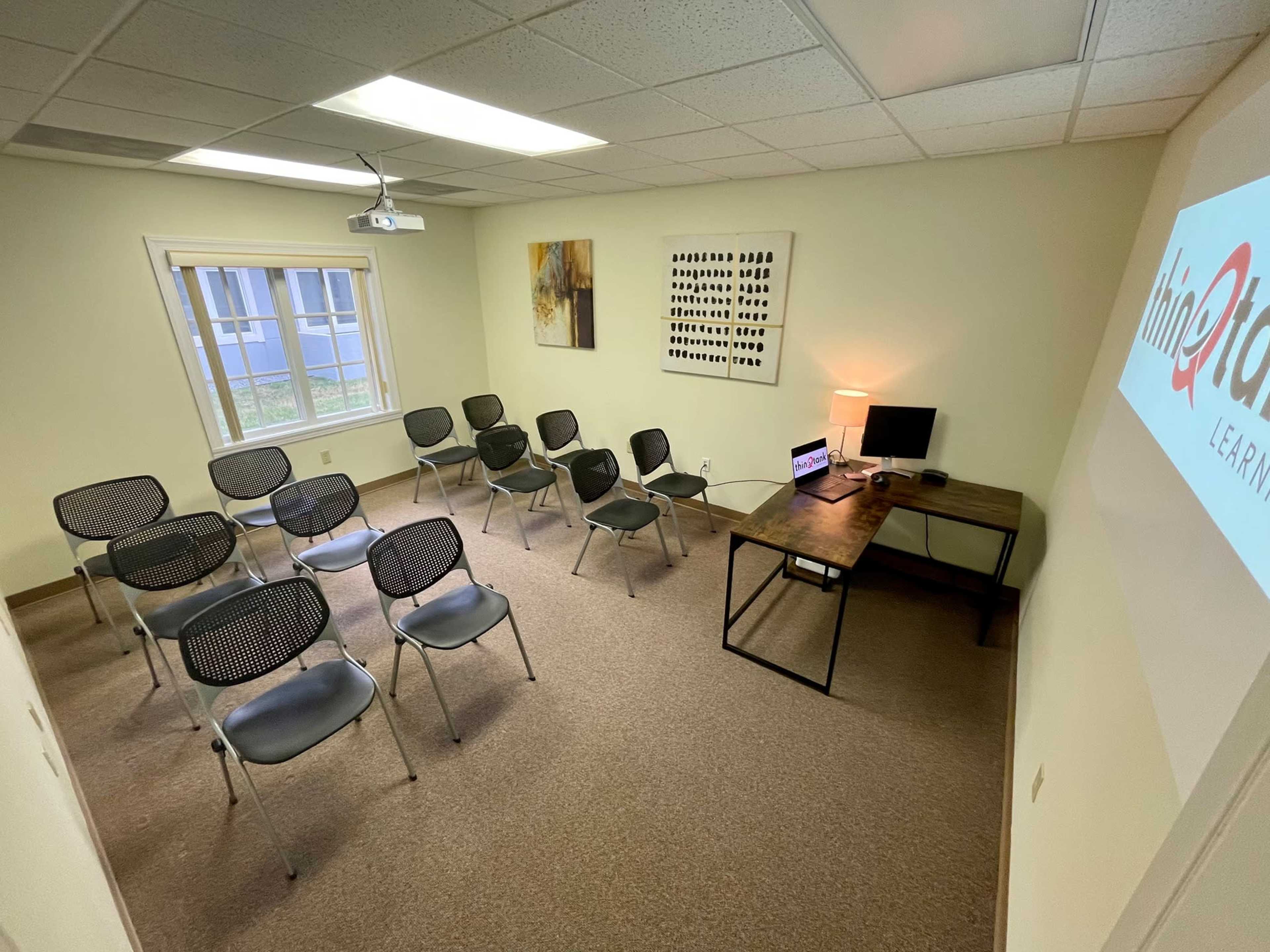 A small meeting room with seated chairs arranged in rows, a desk with a laptop, and a wall-mounted projector displaying a logo.