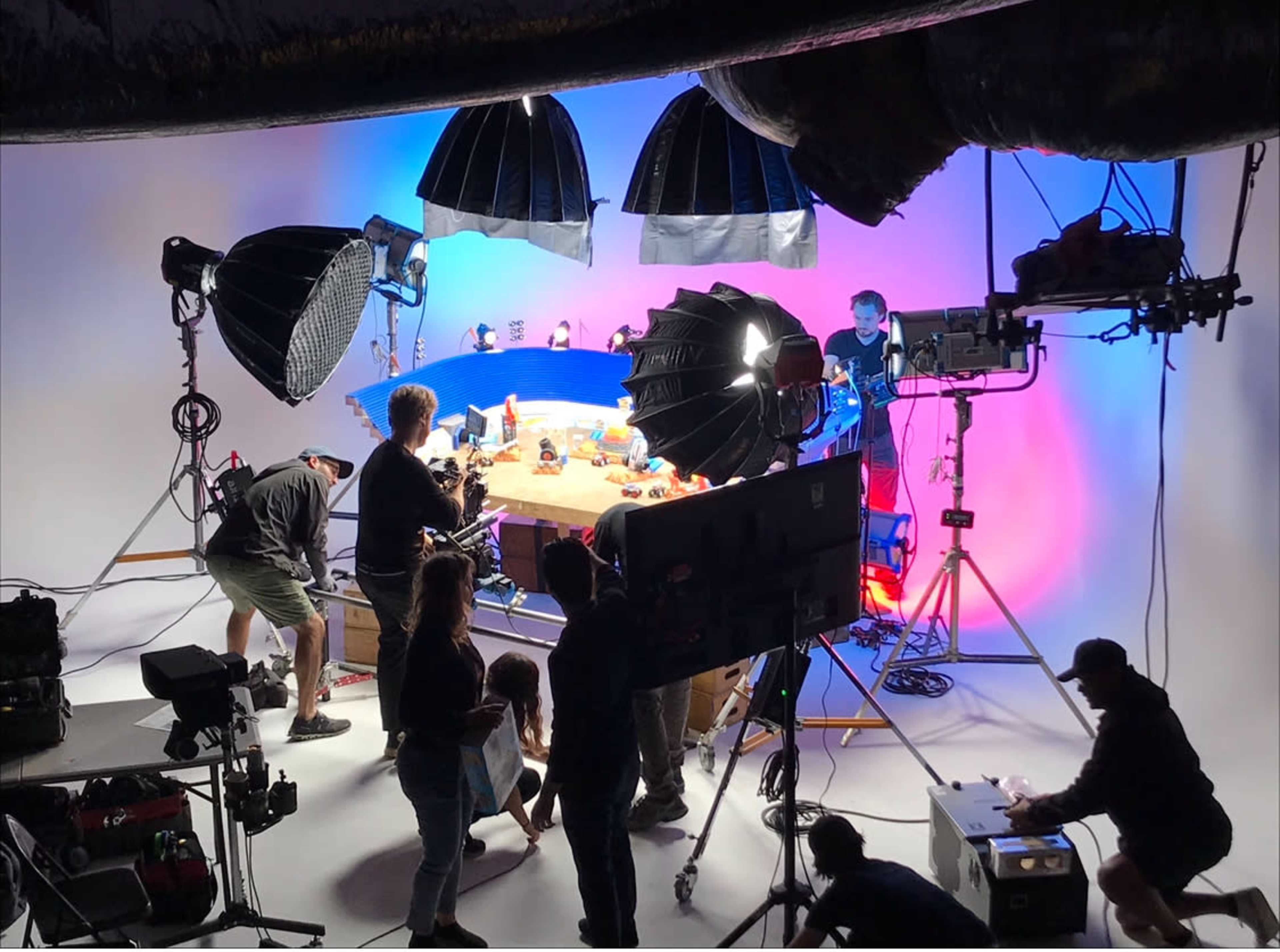 A film crew is set up in a studio, with various lighting equipment and cameras positioned around a table displaying props.