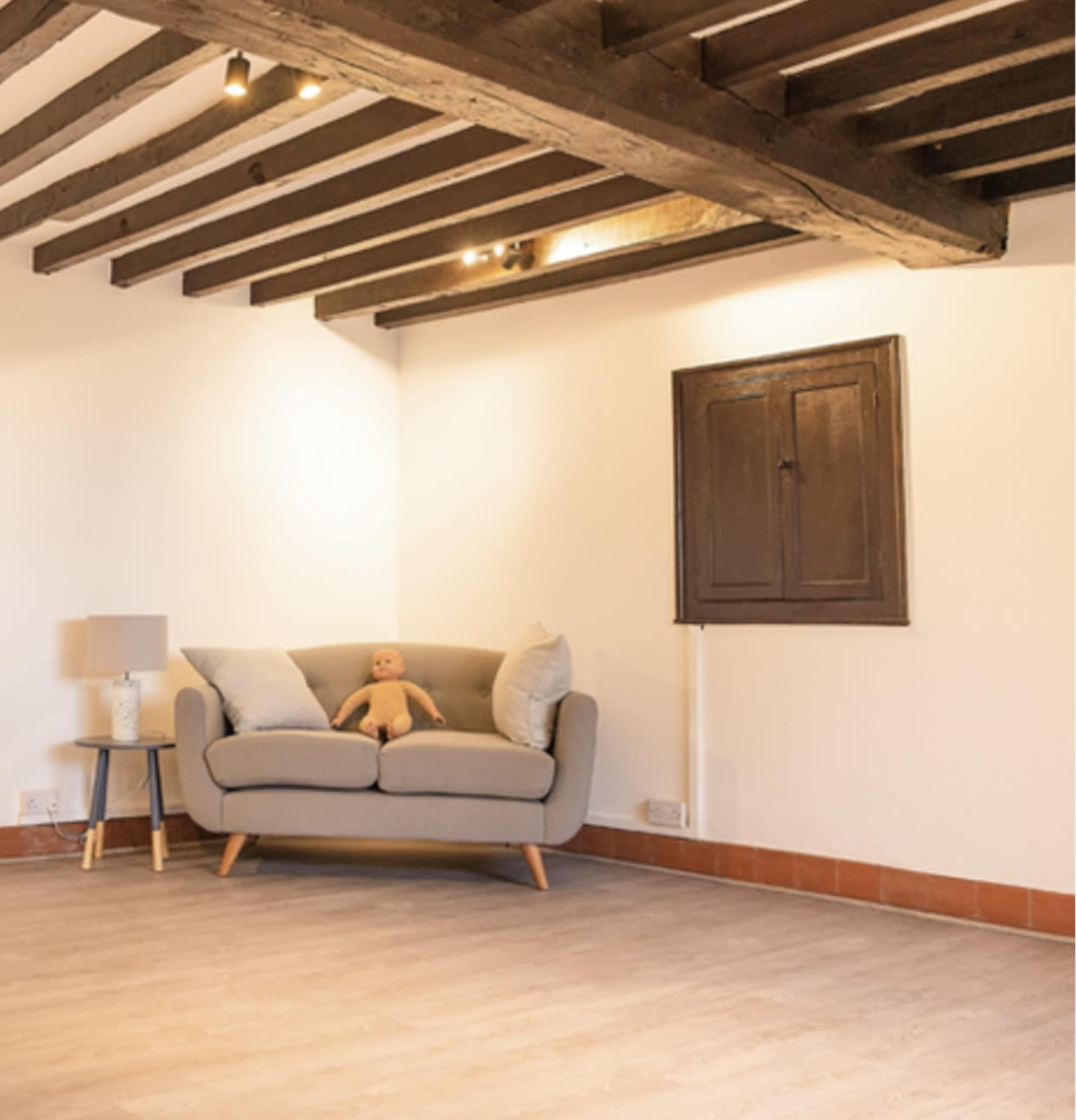 A gray sofa with decorative pillows and a doll sits in a sparsely furnished room featuring wooden beams and a small window.