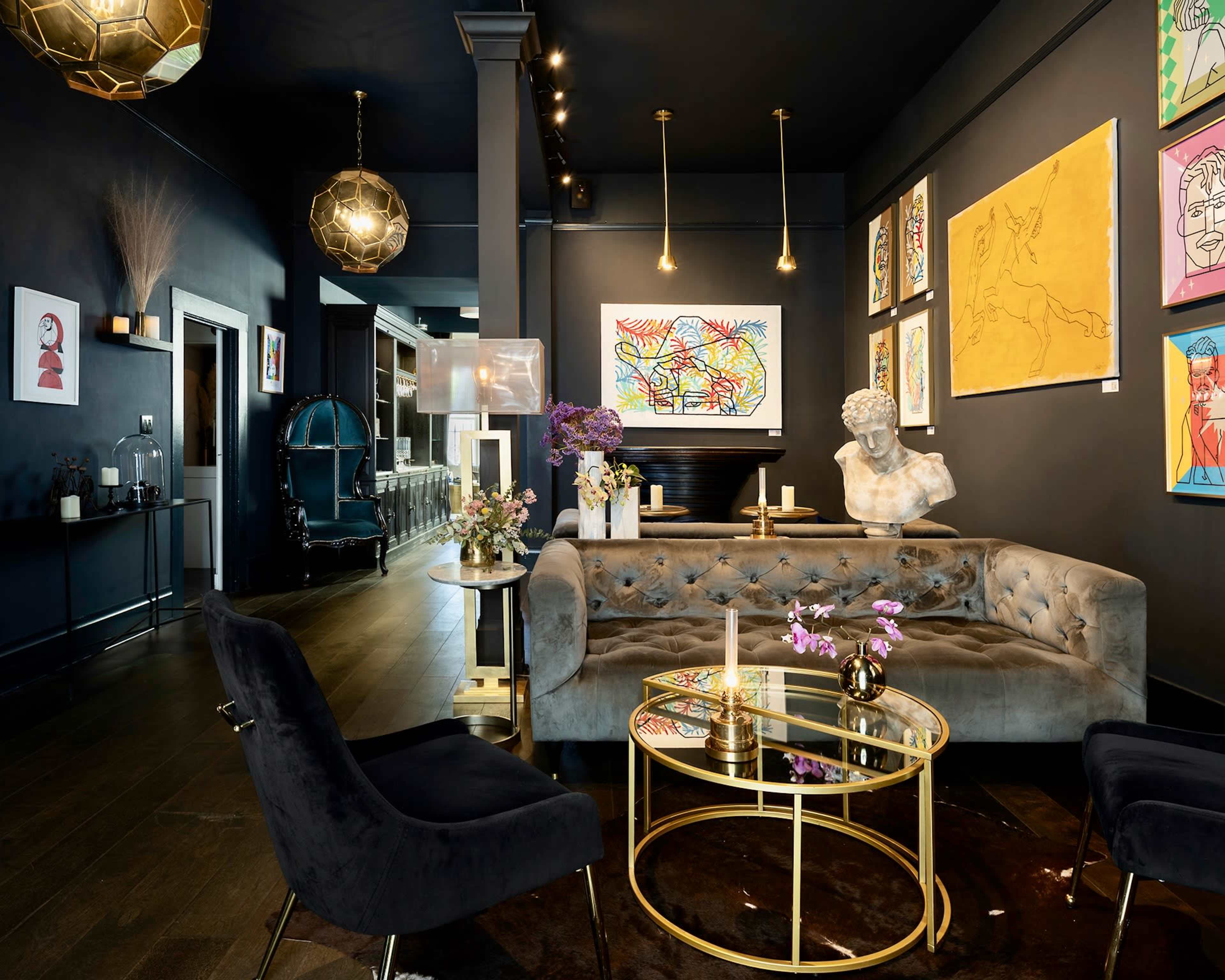 A modern lounge features a gray velvet sofa, a glass coffee table, and colorful artworks on dark walls, with stylish lighting fixtures above.