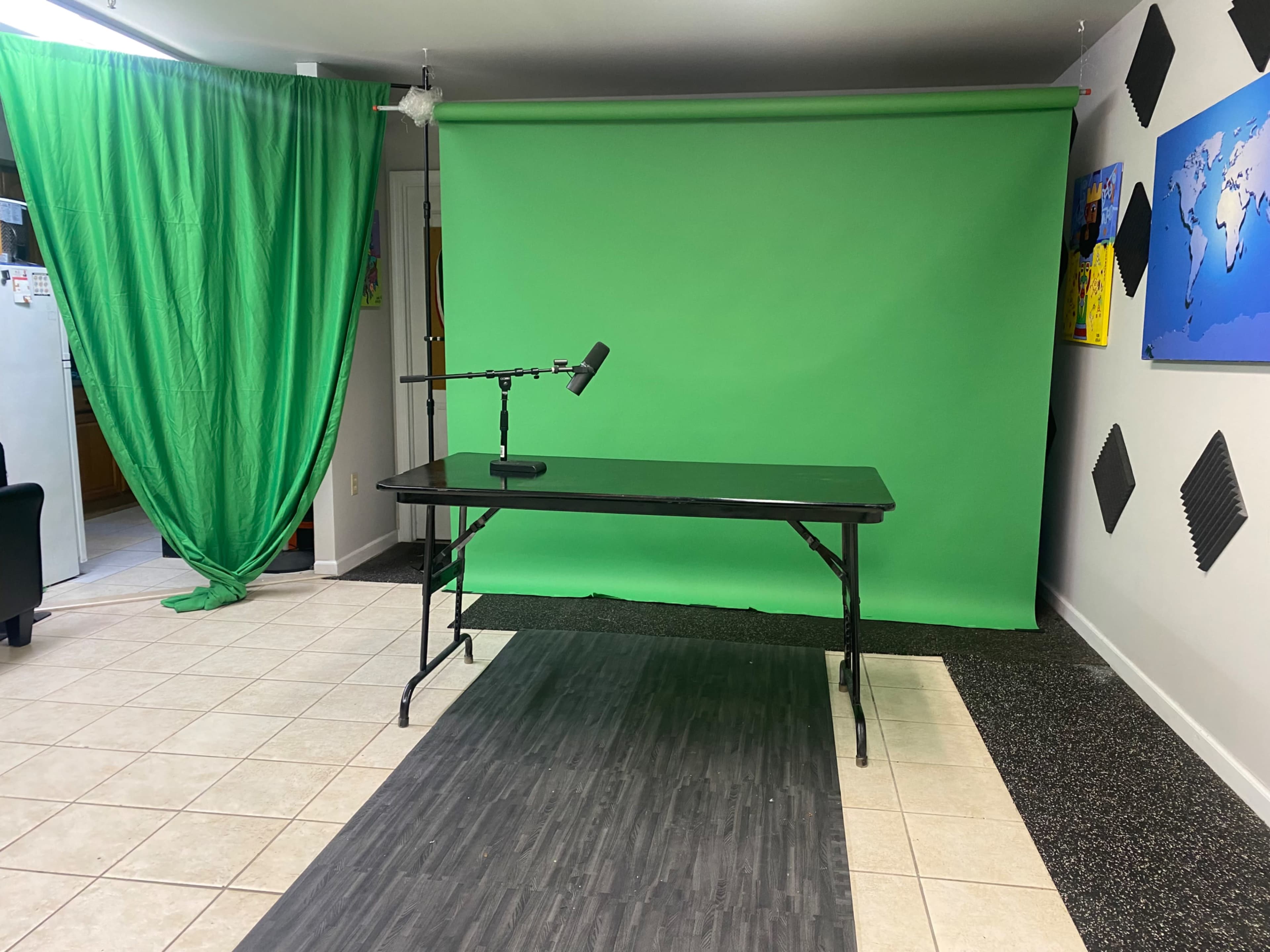 A room set up for video production, featuring a green screen, a black tabletop, a microphone on a stand, and green curtains.
