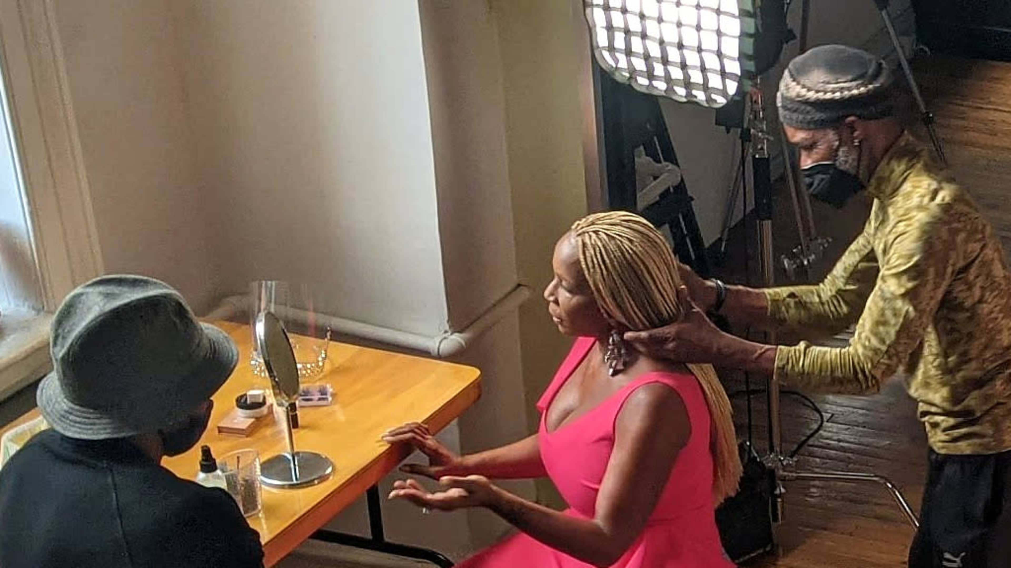 A woman in a pink dress sits at a table with a mirror as two individuals help style her hair under bright lighting.
