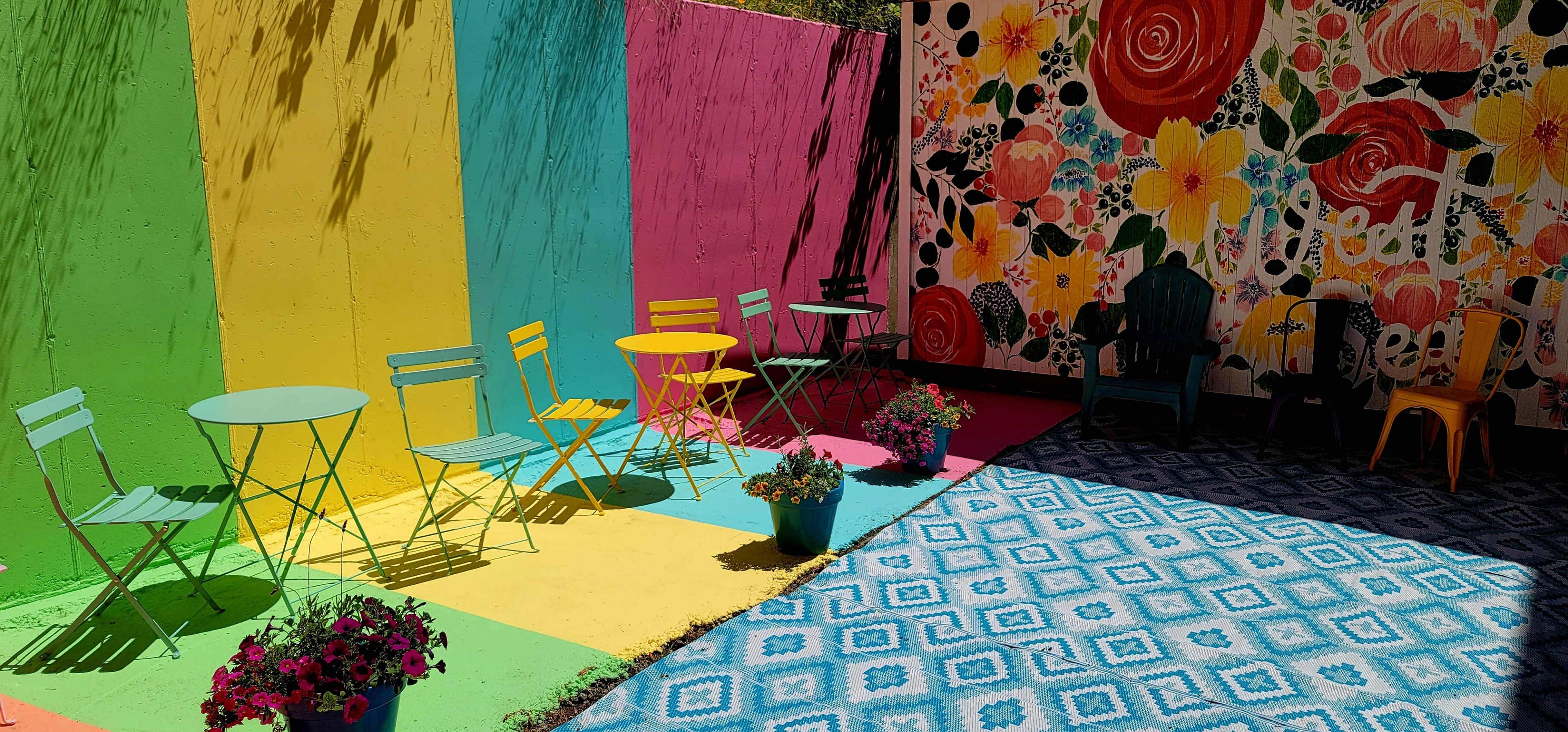 The scene depicts a vibrant outdoor seating area with colorful chairs and tables surrounded by a mural of flowers and a geometric patterned rug.
