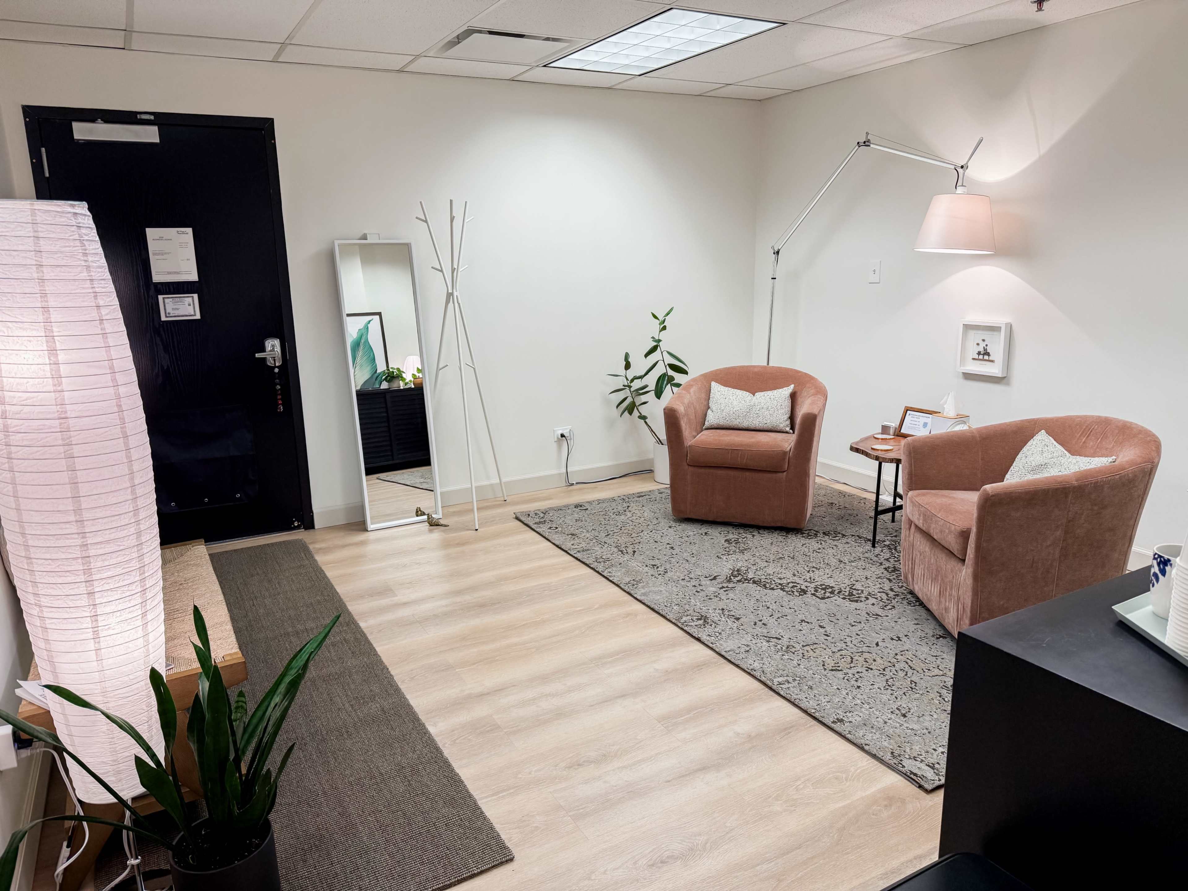 The image shows a neatly arranged office space featuring two chairs, a floor lamp, a mirror, and a plant, with light wood flooring and a decorative rug.