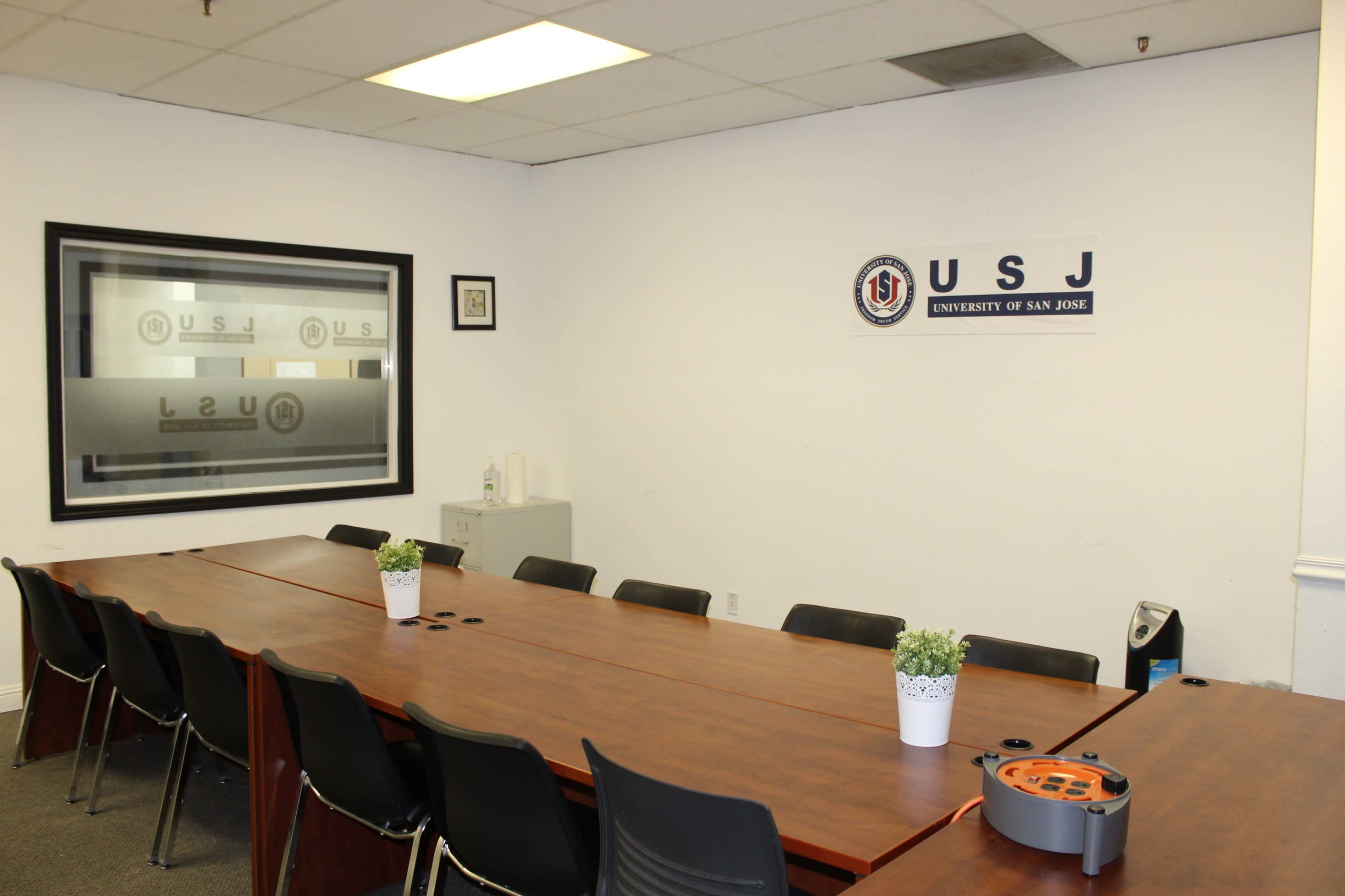 A large conference room with a long wooden table, black chairs, and a wall displaying the University of San Jose logo.